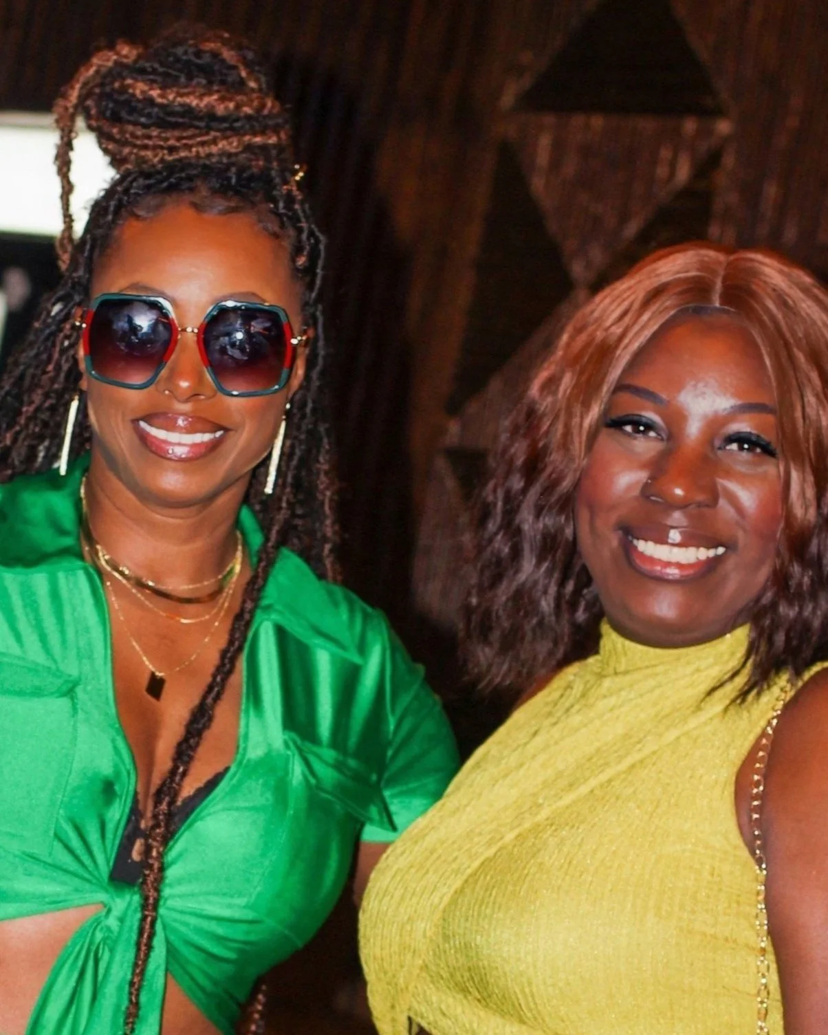 Two women smiling, one wearing green with sunglasses and braided hair, the other wearing yellow with wavy hair, indoors.