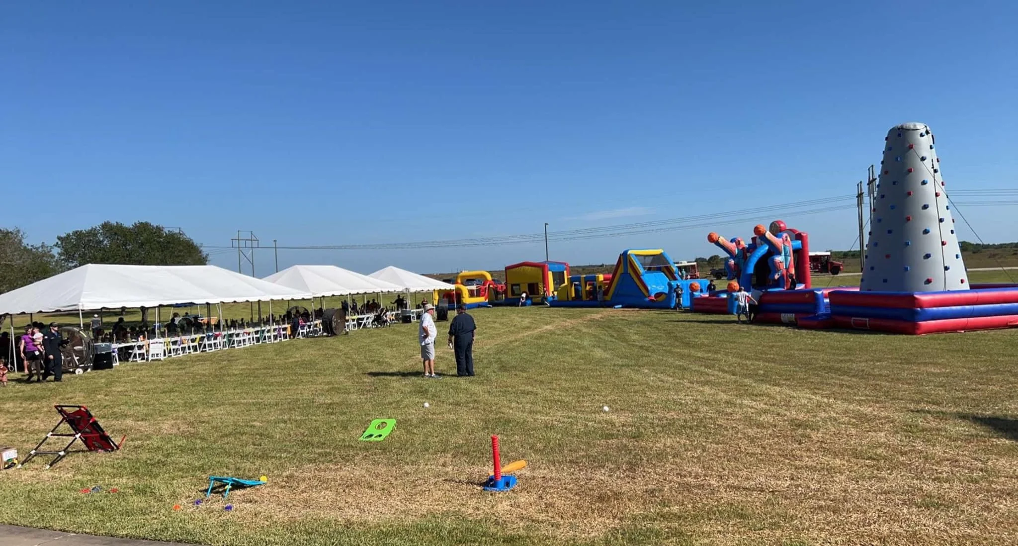 A large outdoor field with inflatable play structures and tents, including a climbing wall, bouncy castles, and a tent with tables and chairs, under a clear blue sky.