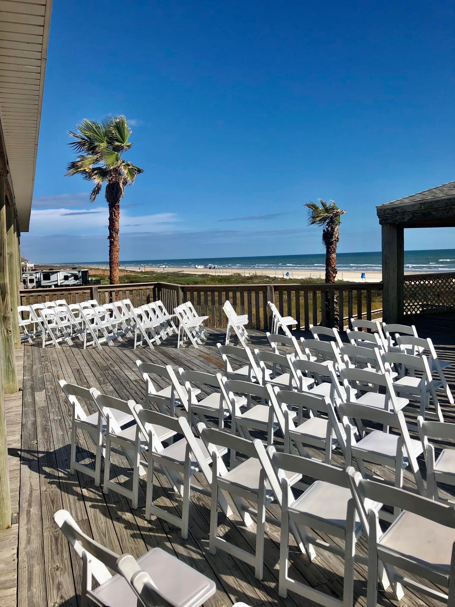 Empty outdoor wooden deck with white folding chairs facing the beach, with palm trees and ocean in the background, under a bright blue sky.