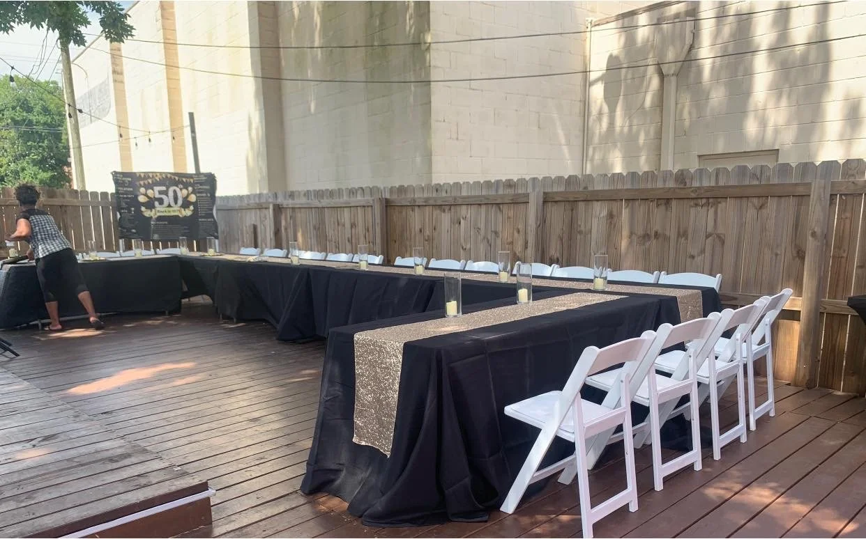 Outdoor wooden deck party setup with black tablecloths, white chairs, and a large sign celebrating 50th birthday against a wooden fence.