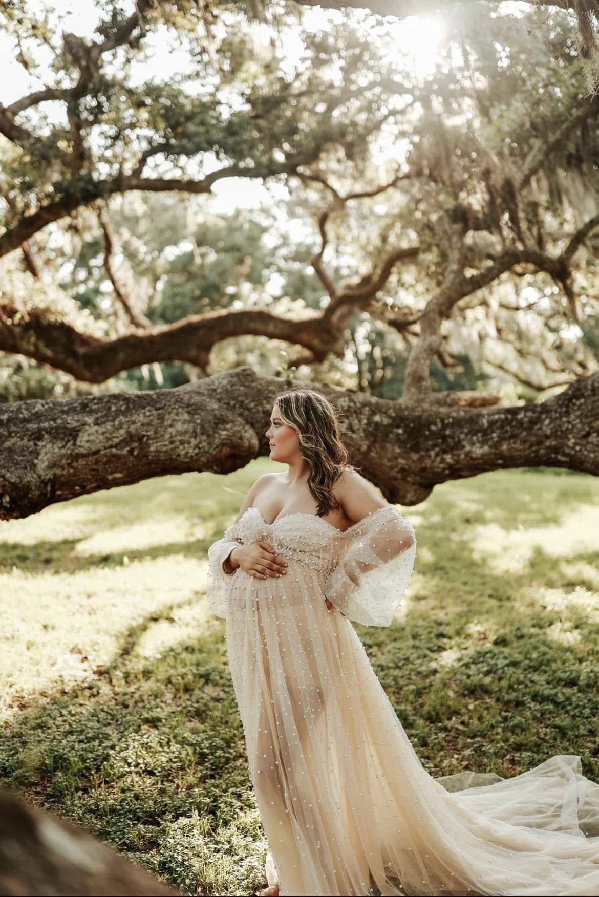 A bride in a flowing, off-the-shoulder dress standing outdoors under a large, sprawling tree, with sunlight filtering through the branches.
