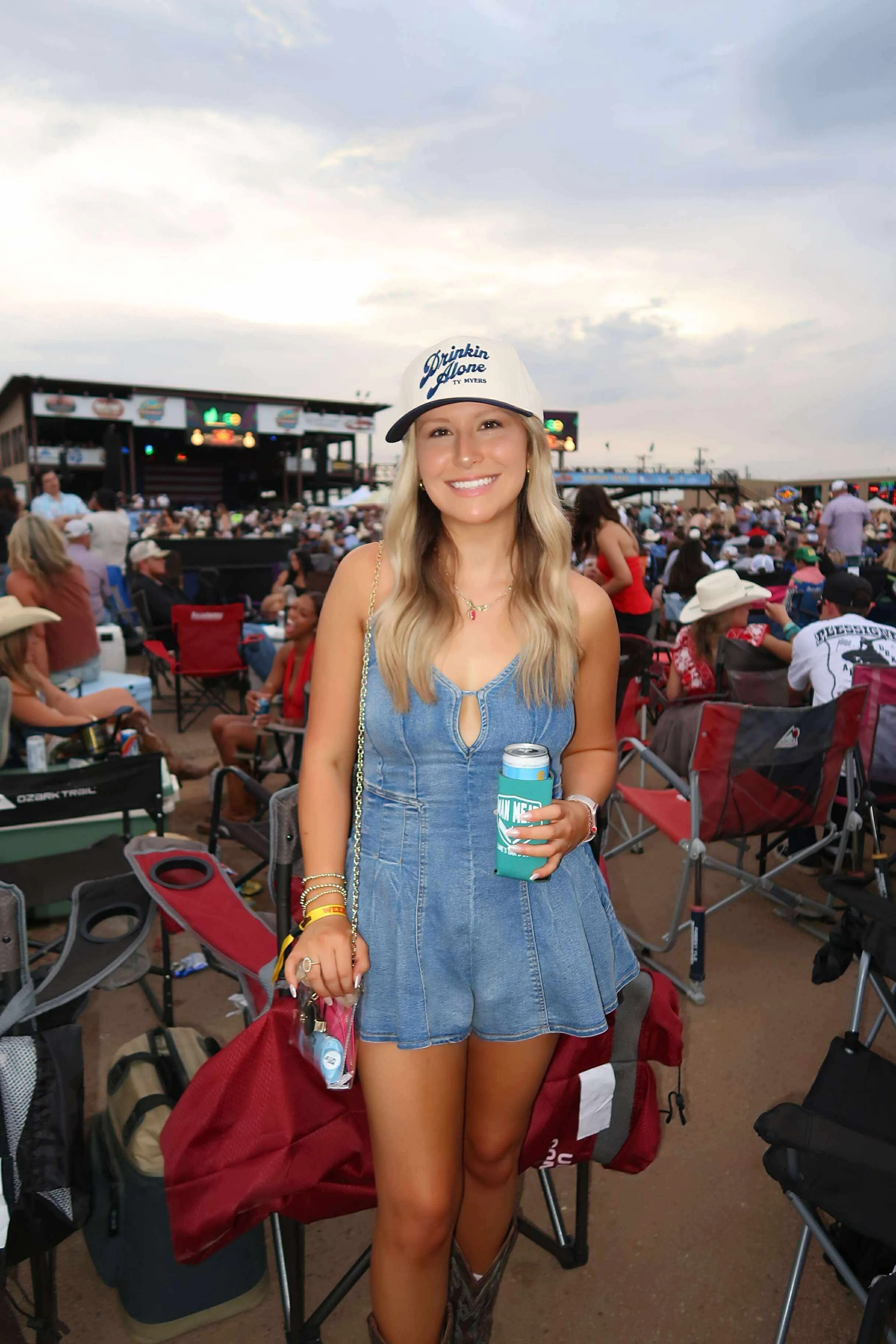 A young woman smiling, wearing a gray baseball cap with 'Drinkin Alone TV Myers' written on it, a denim romper, and holding a can, at an outdoor concert or festival with many people sitting and standing in the background.