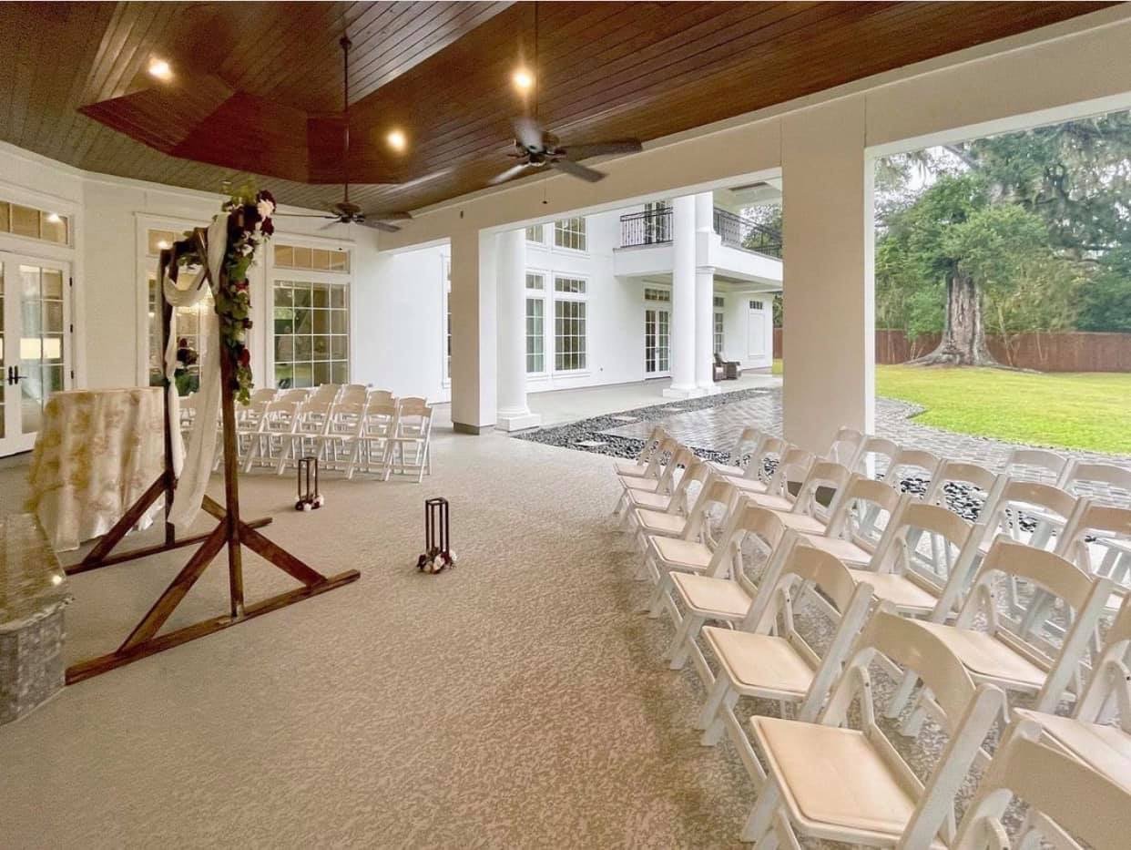 Indoor wedding ceremony setup with white chairs arranged facing an arch decorated with flowers, inside a spacious covered patio area with large open sides, next to a lawn and large tree.