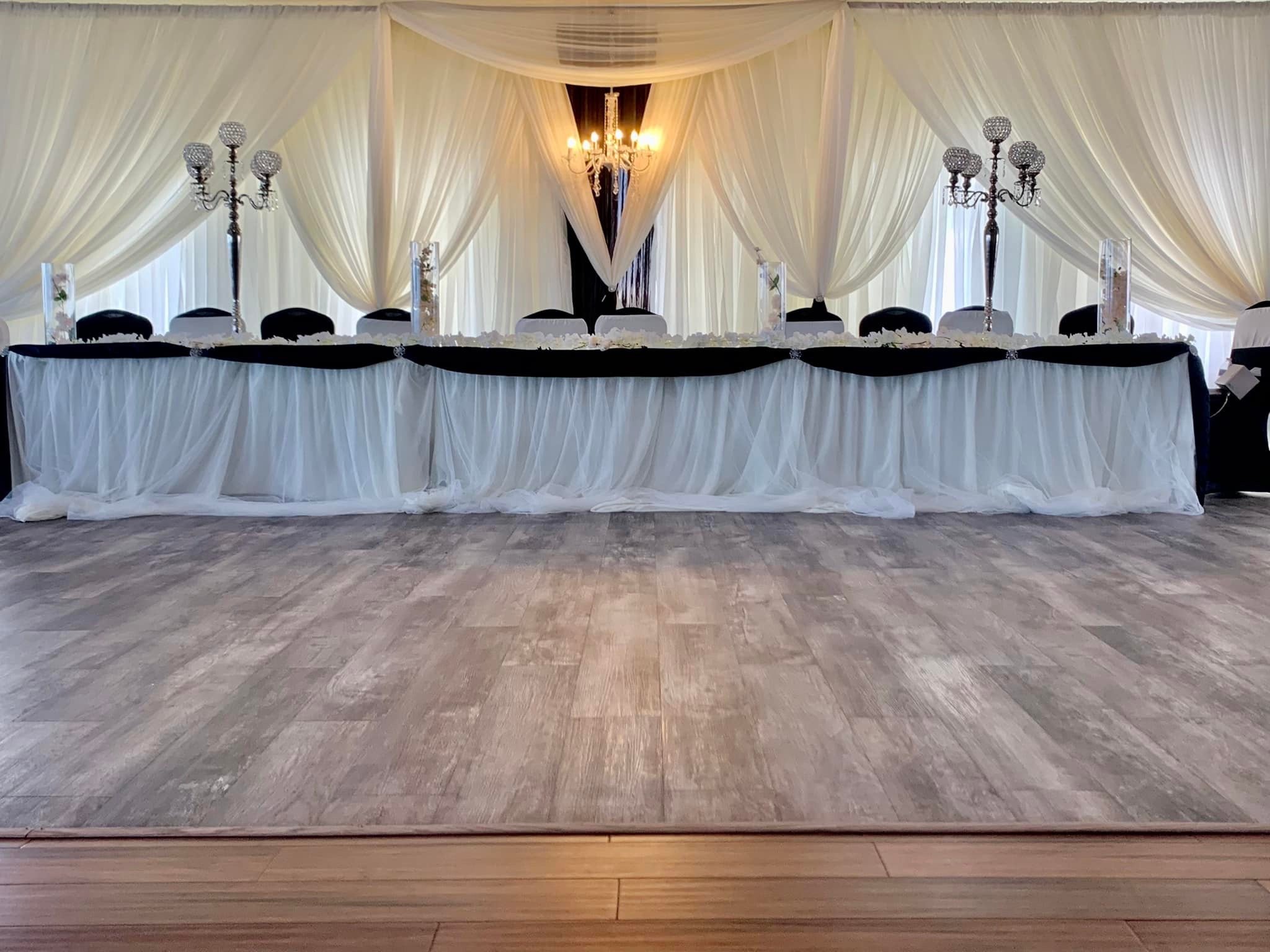 Elegant wedding reception table decorated with white and black drapes, crystal chandeliers, candelabras, and floral centerpieces, set against a backdrop of white curtains.