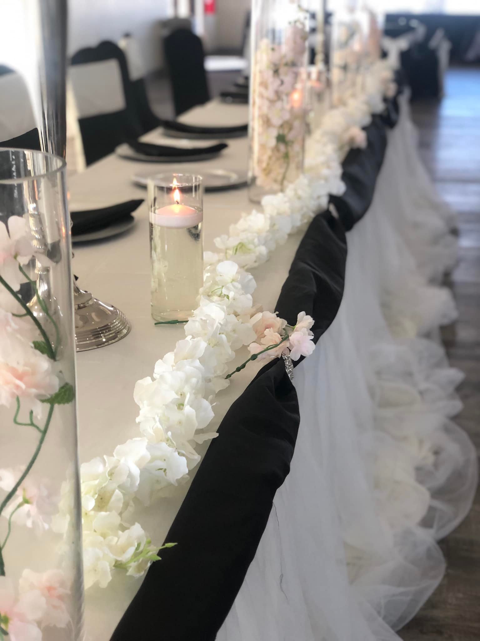 Elegant banquet table decorated with white and pink flowers, black and white napkins, and a tall candle in a glass holder, set for a formal event.