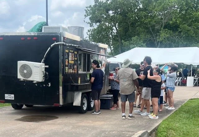 People standing in line at a black food truck with a white tent in the background.