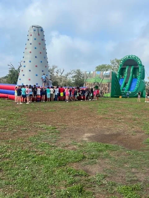 Group of children and adults at a fairground with an indoor rock climbing wall and a large inflatable slide in the background.