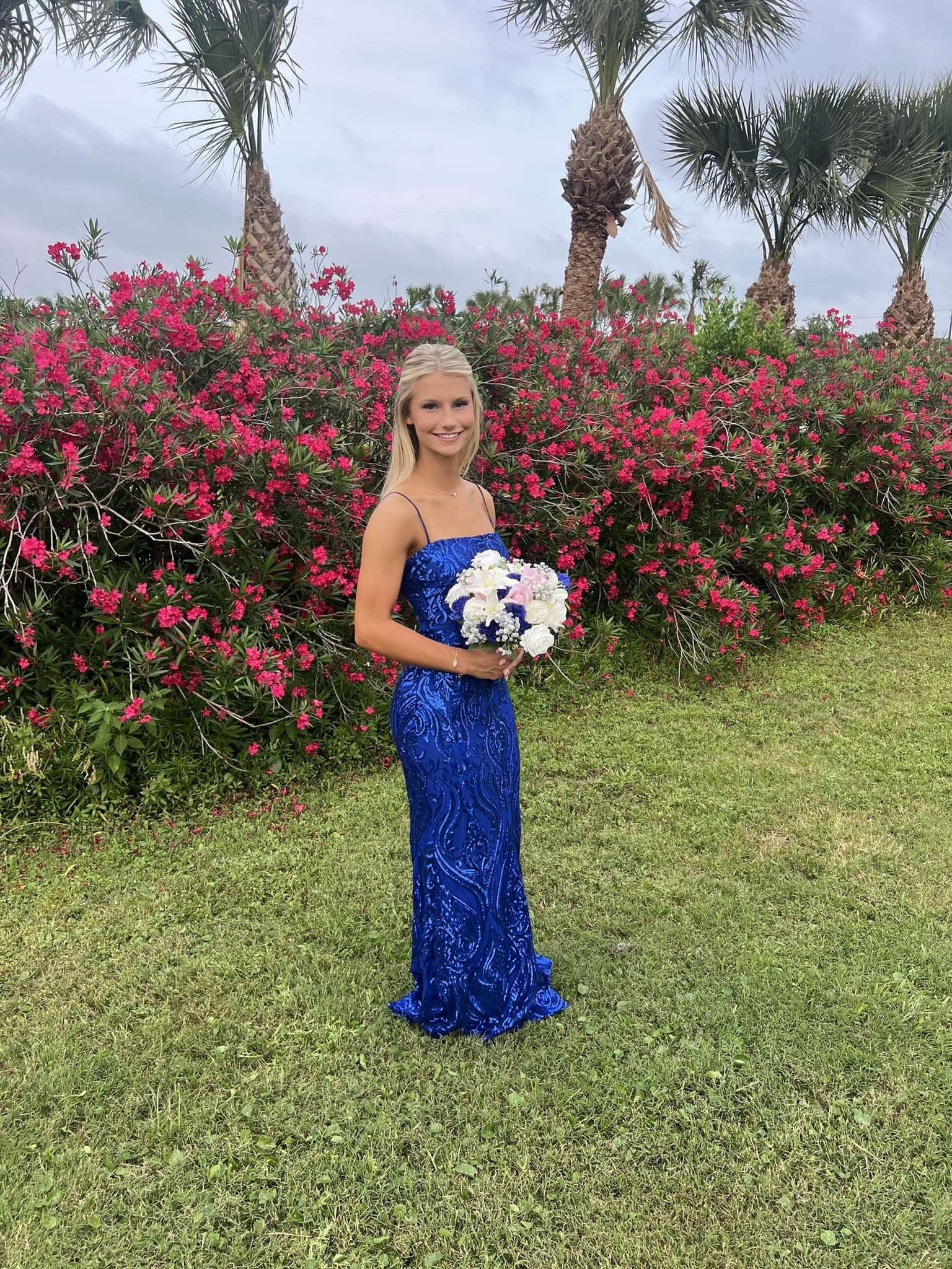 A young woman in a blue dress holding a bouquet of flowers outside in front of pink flowering bushes and palm trees.