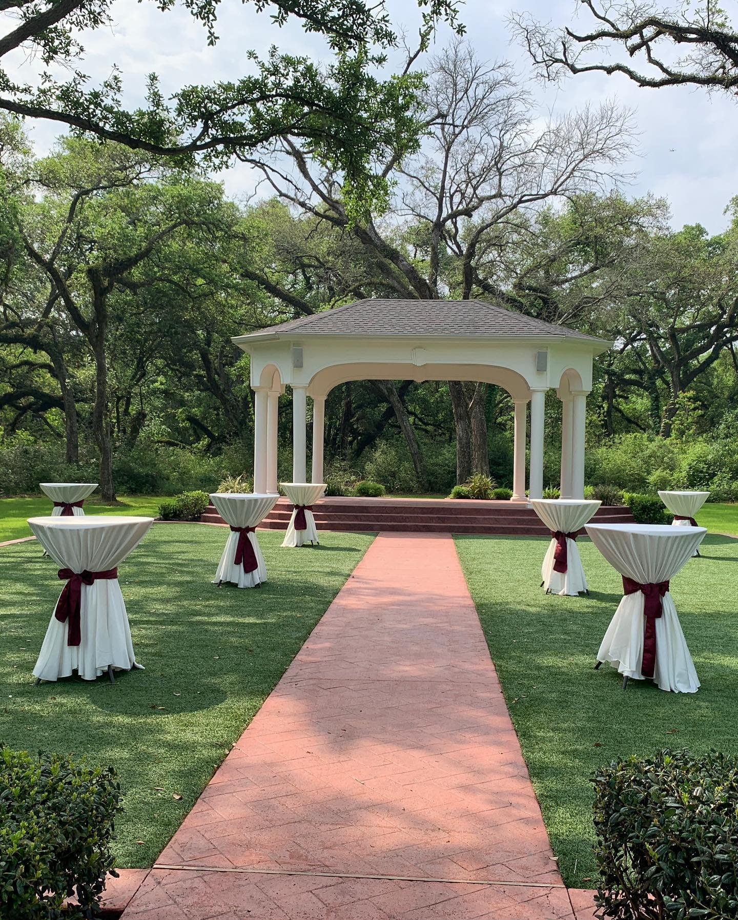An outdoor wedding or event space with a white gazebo, surrounded by trees, with tall cocktail tables draped in white cloth and maroon ribbons lining a brick pathway leading to the gazebo.
