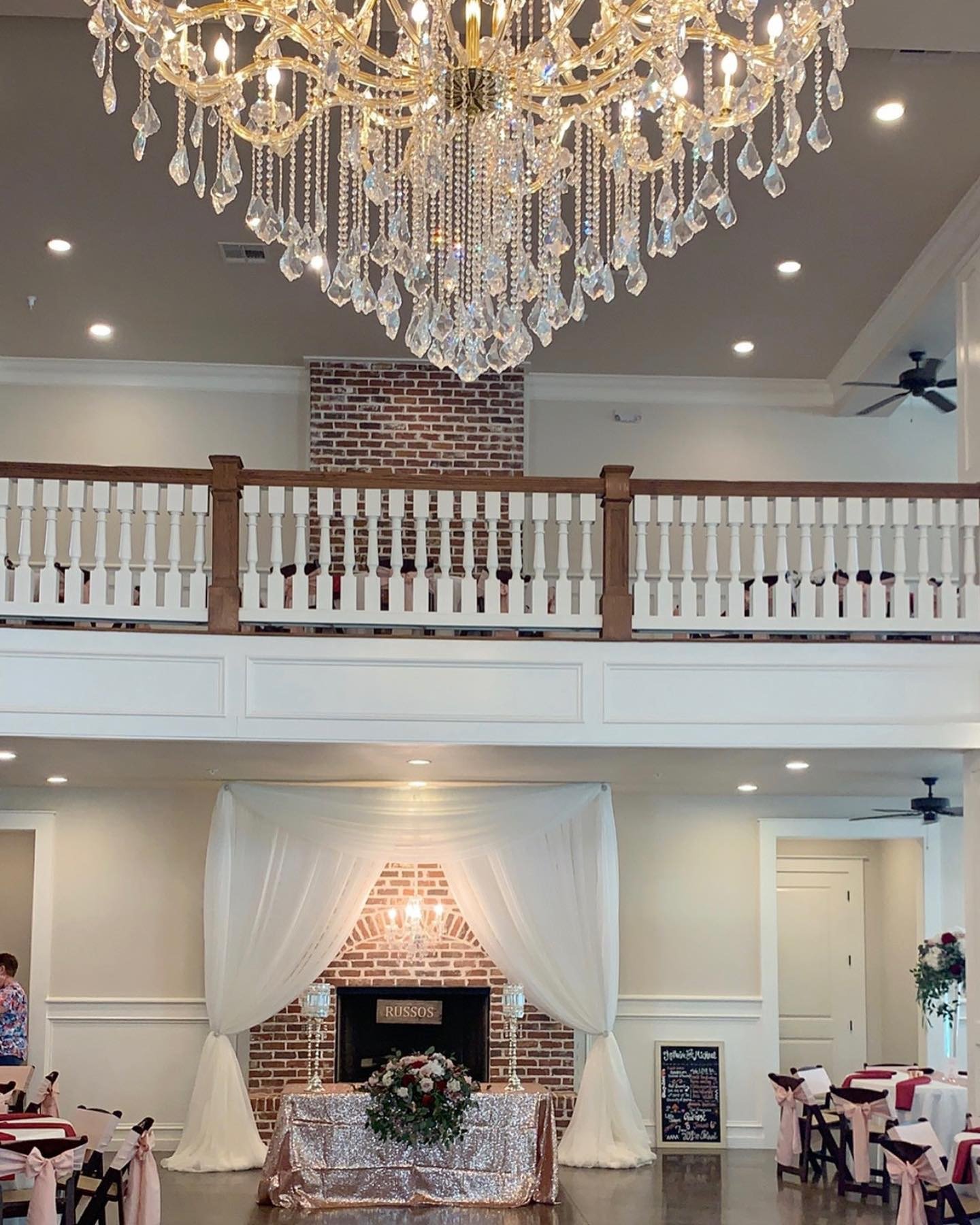 An elegant event space decorated with a large crystal chandelier hanging from the ceiling, a brick fireplace with a chandelier above a table in front of white drapes, and round tables with pink bows on the chairs.