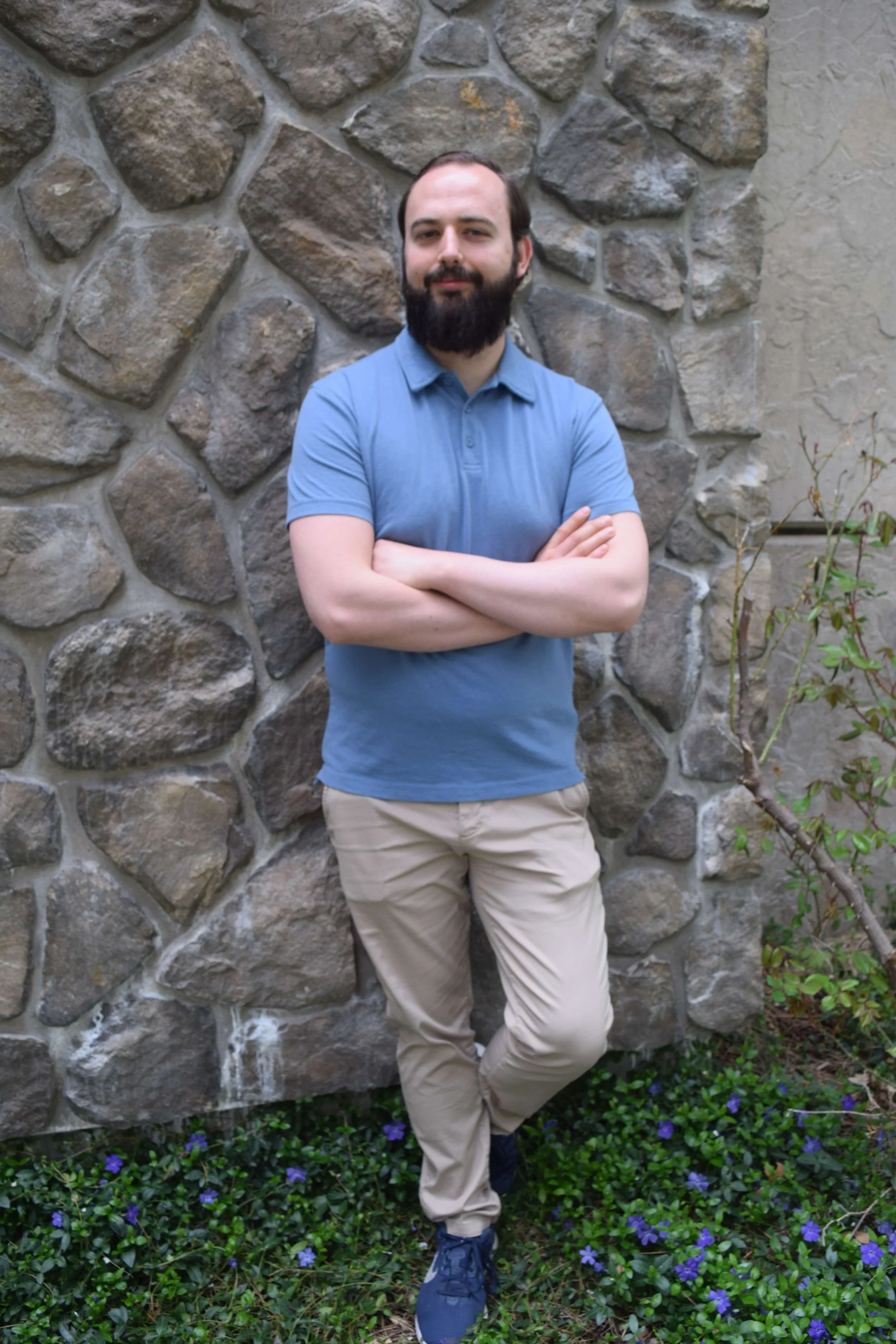 Photo of Taylor Wood, owner of Chariot Bookkeeping, wearing a blue polo shirt and khaki pants, stands with crossed arms in front of a stone wall, with green plants and small purple flowers at his feet.