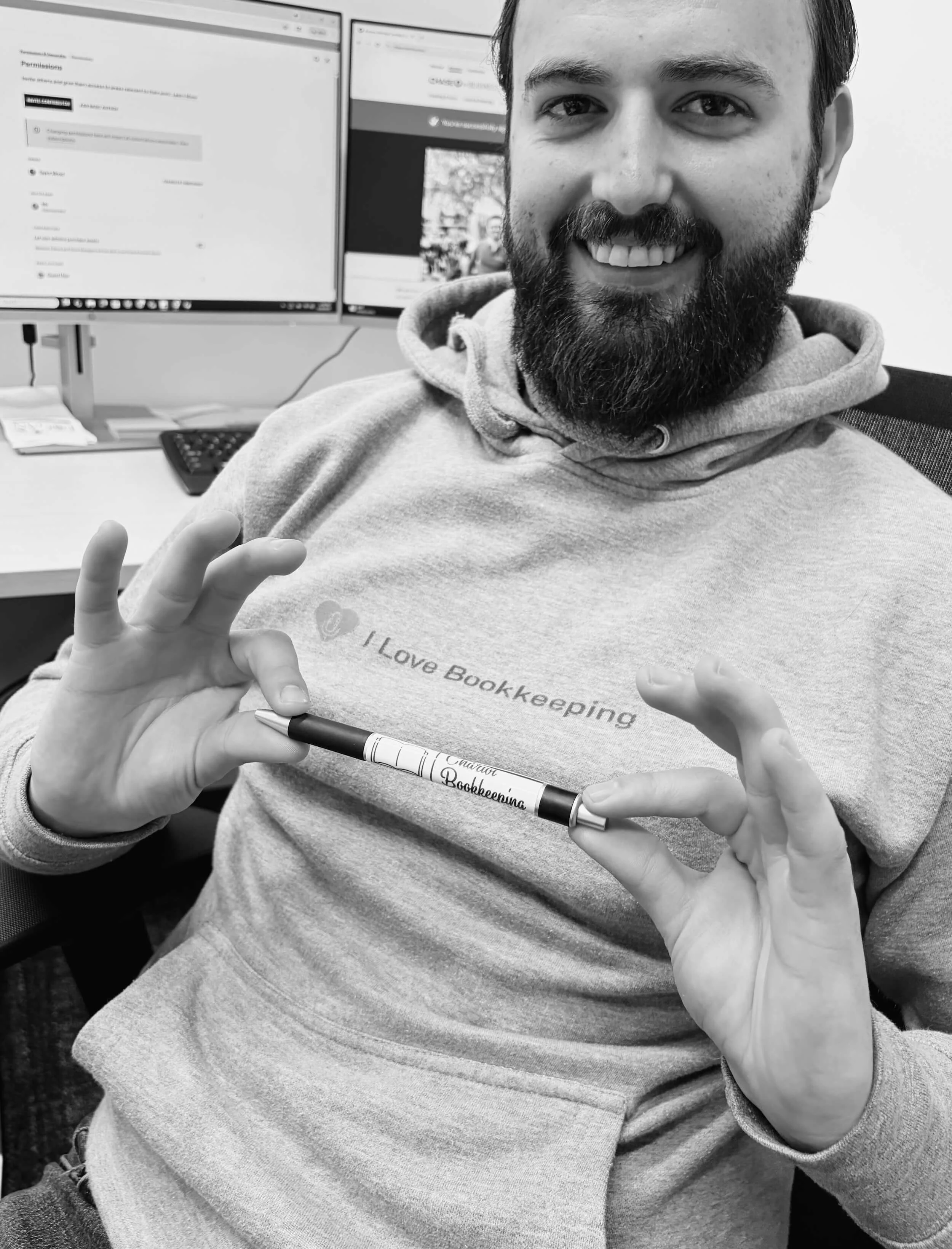 Taylor Wood smiling at the camera, holding a pen that says 'Chariot Bookkeeping,' while sitting in front of dual computer monitors, wearing a hoodie with the text 'I Love Bookkeeping'.