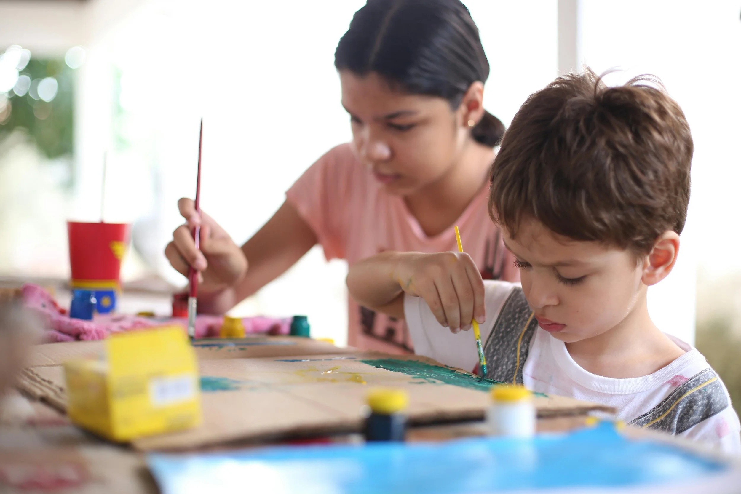 A young girl and a boy painting together at a table with paintbrushes and colorful paints.