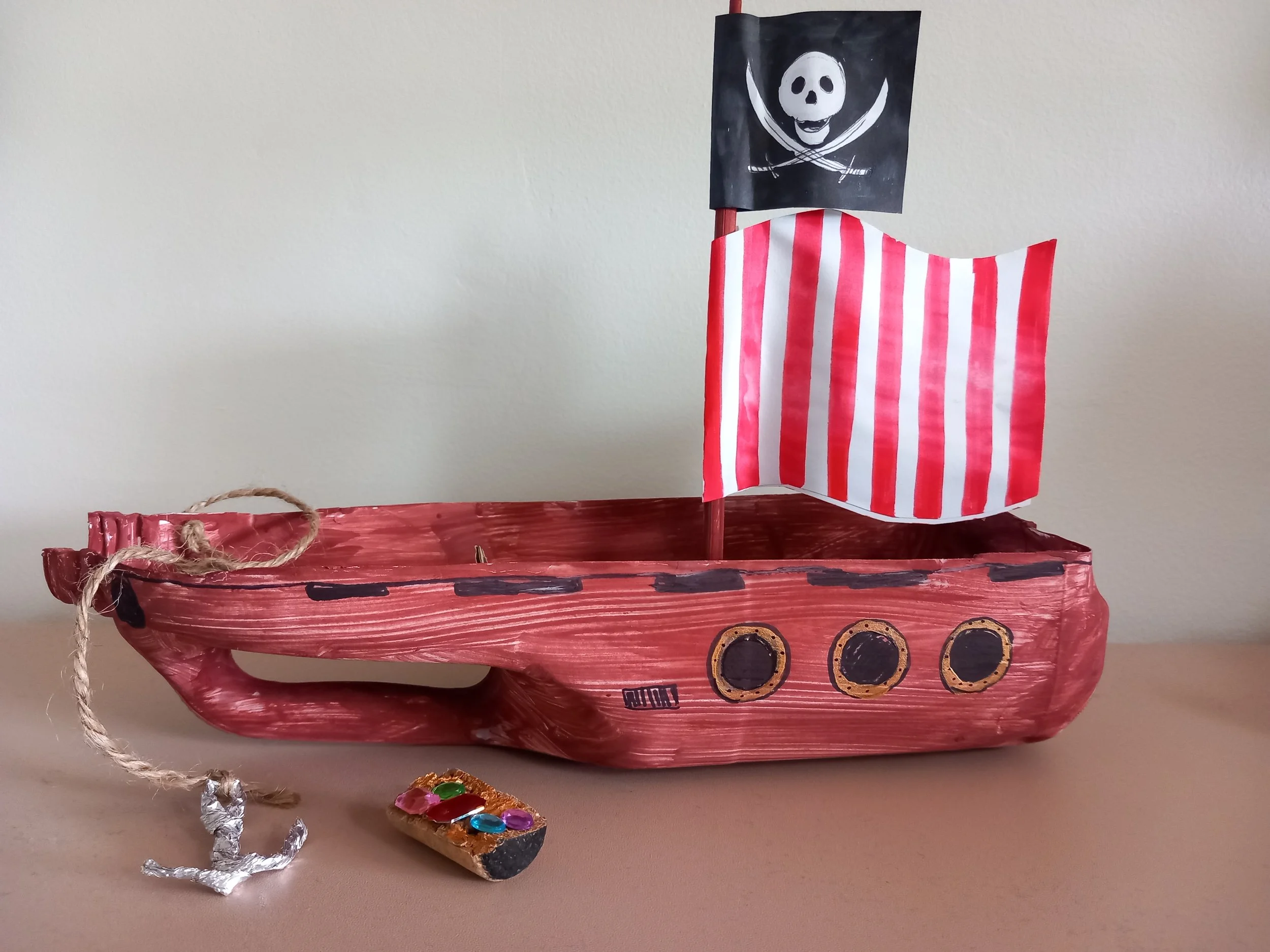 Handmade pirate ship craft with red body, black and gold portholes, red and white striped sail, and black skull and crossbones flag, with a small anchor made from foil, string, and decorated with colorful stones.
