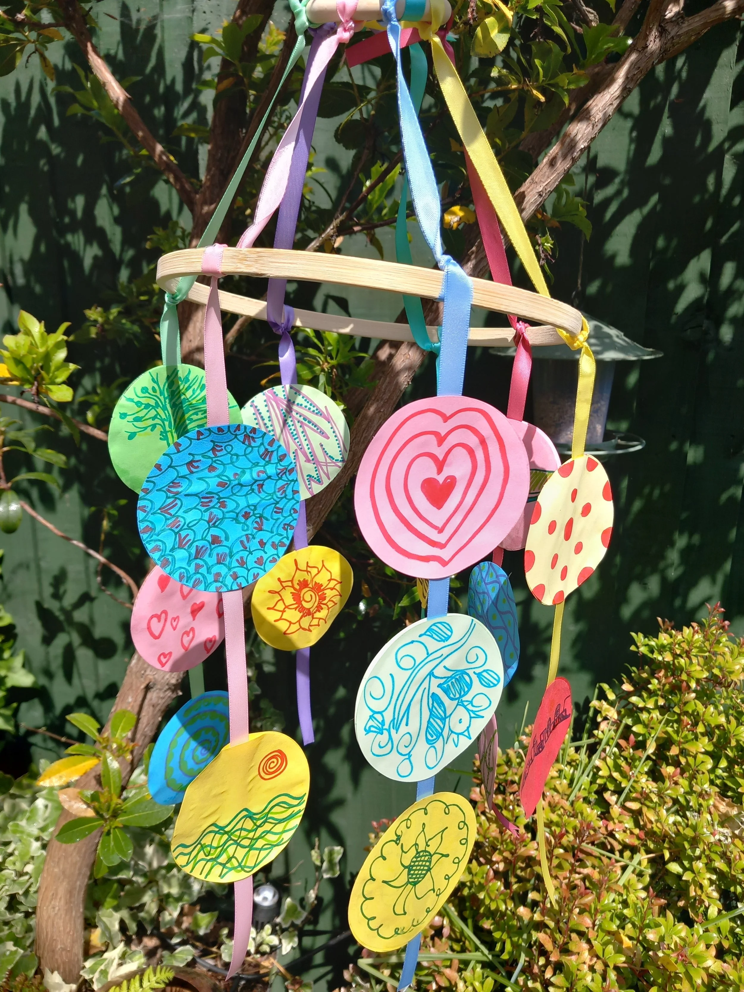 Colorful paper circles with drawings hanging on ribbons from a wooden hanger, outdoors among green plants.
