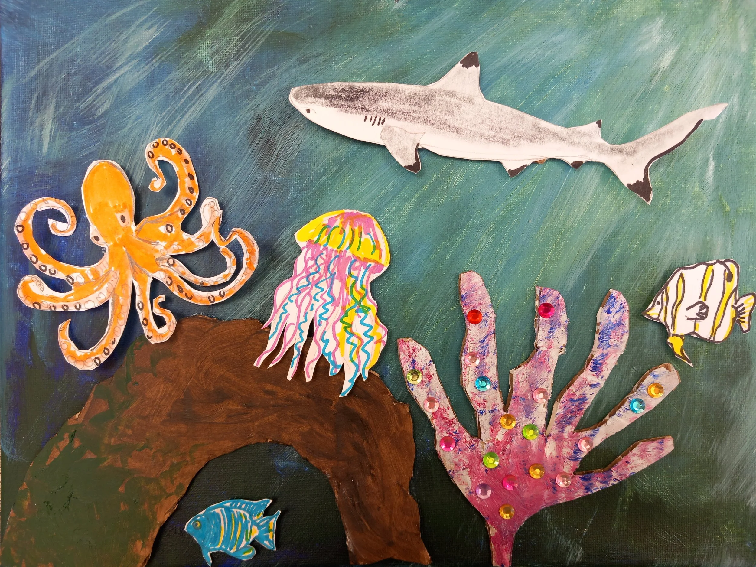 Underwater scene with a paper-crafted octopus, fish, jellyfish, shark, and coral against a painted blue-green background.