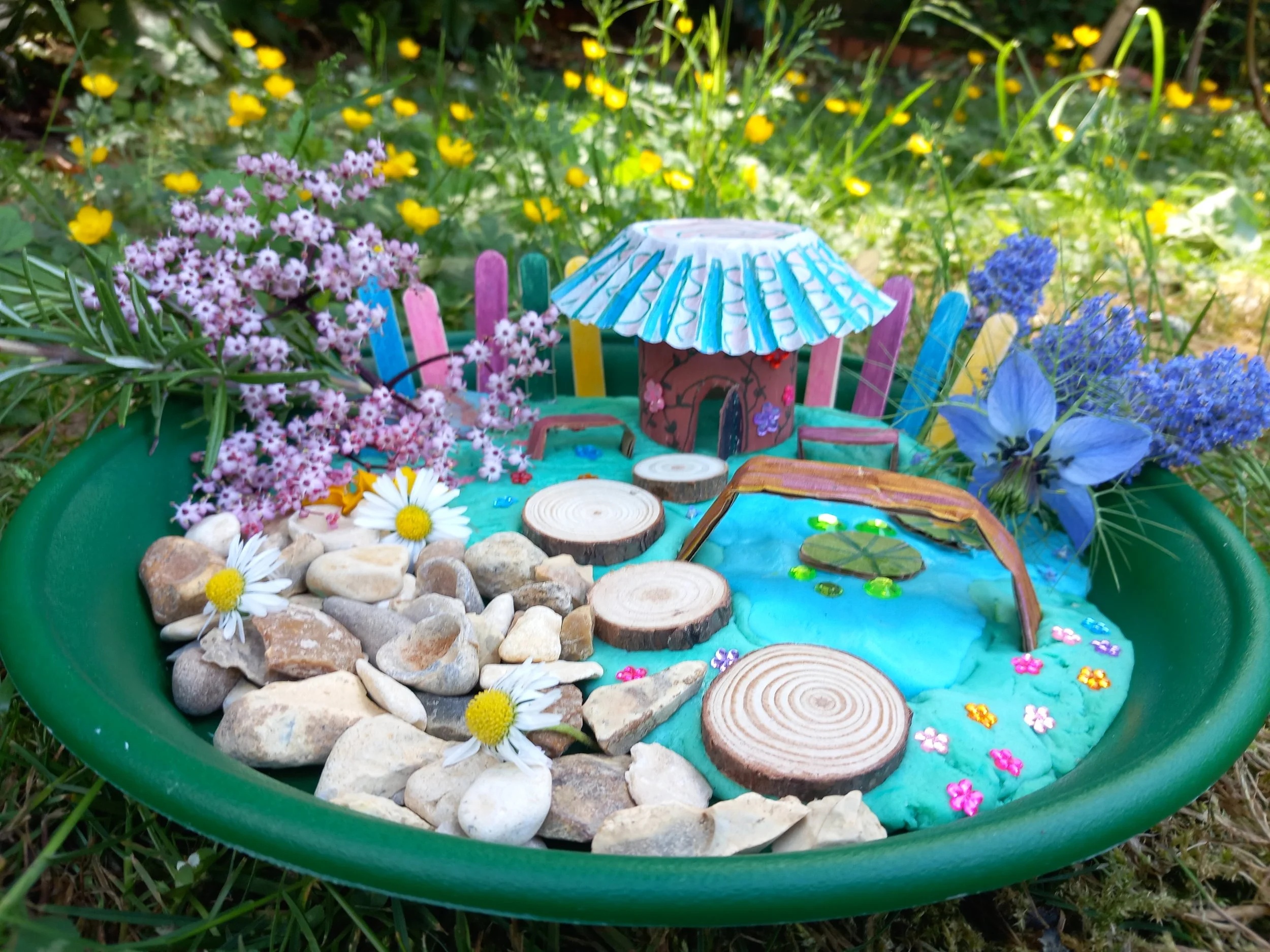 Miniature garden scene with colorful handmade paper elements, small rocks, artificial flowers, and a painted paper umbrella inside a green tray.