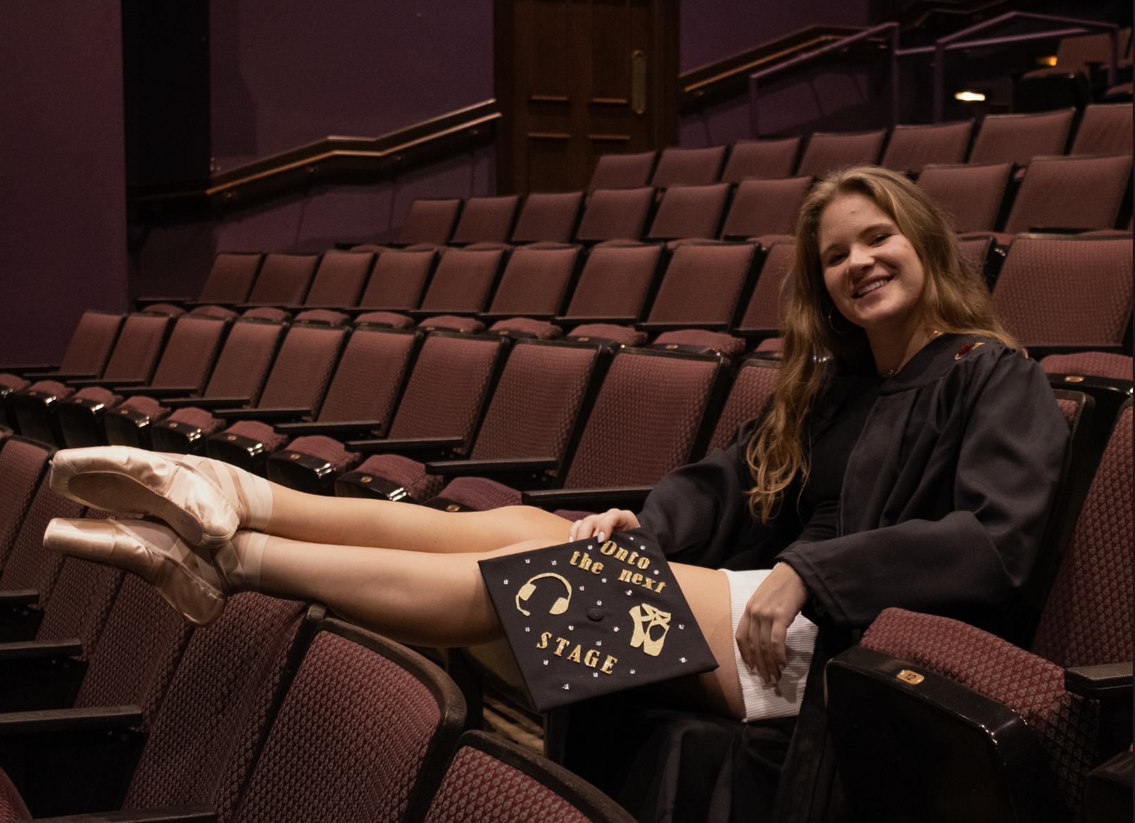 A young woman in a graduation gown and cap sitting in an empty theater, smiling, with her feet up on a theater seat armrest, holding a diploma that says 'On to the next stage' and has gold stars and a logo.