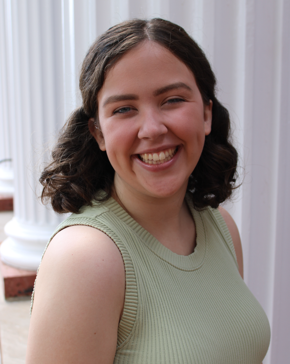 A young woman with curly brown hair smiling outdoors near white columns.