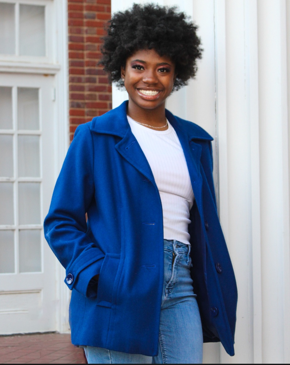 A young Black woman with a big curly hairstyle smiling, wearing a bright blue coat, white shirt, and blue jeans, standing outside near a white door and brick wall.