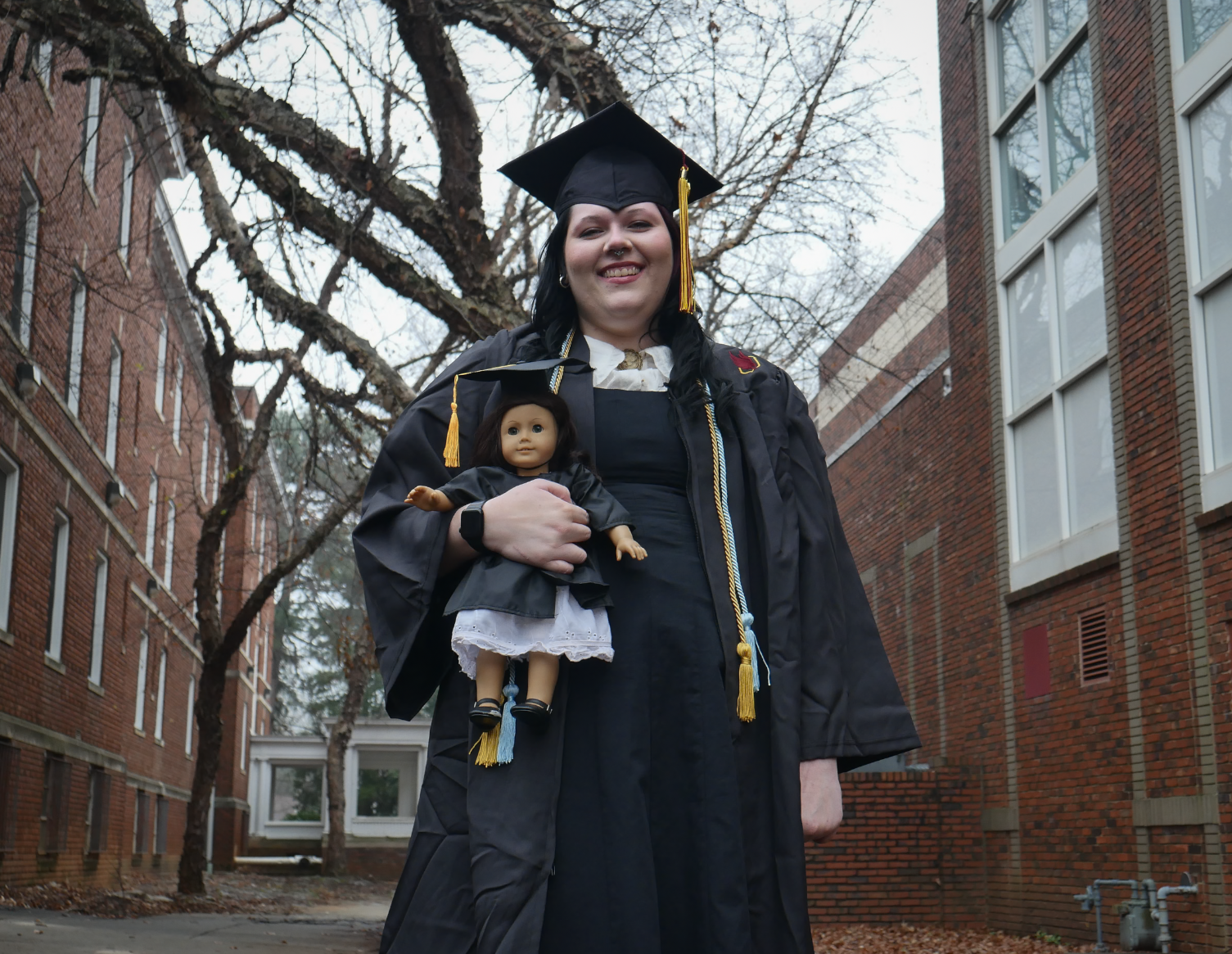 Graduate woman in cap and gown holding a doll dressed as a graduate, outdoors in front of brick buildings and leafless trees.