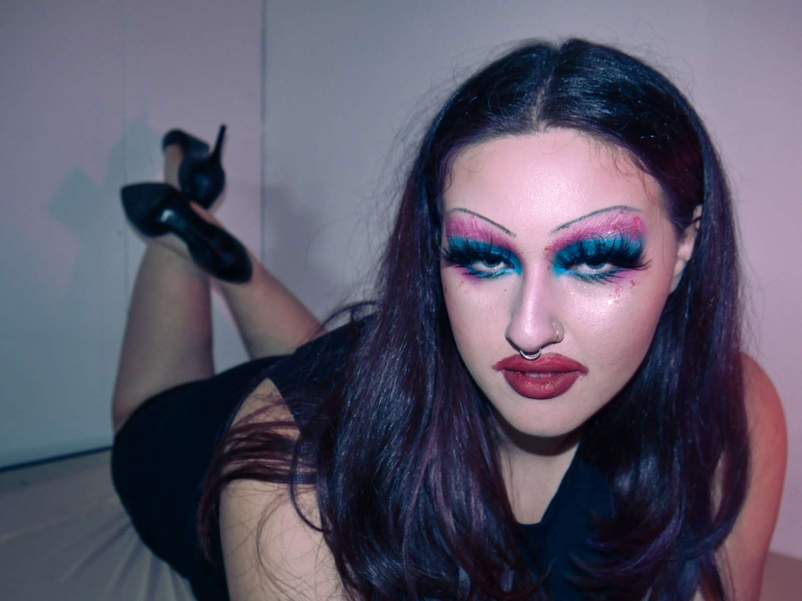 Person with long dark purple hair lying on their stomach on a floor, wearing black high heels and a black outfit, with colorful makeup including blue eyeshadow and glitter, and a septum piercing.
