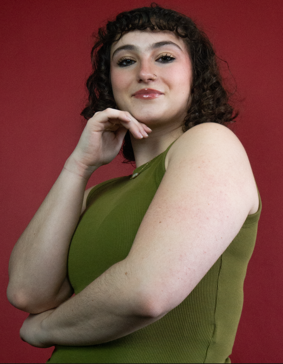 A young woman with curly brown hair and a nose ring, wearing a green tank top, posing with her arm crossed and hand near her chin against a red background.