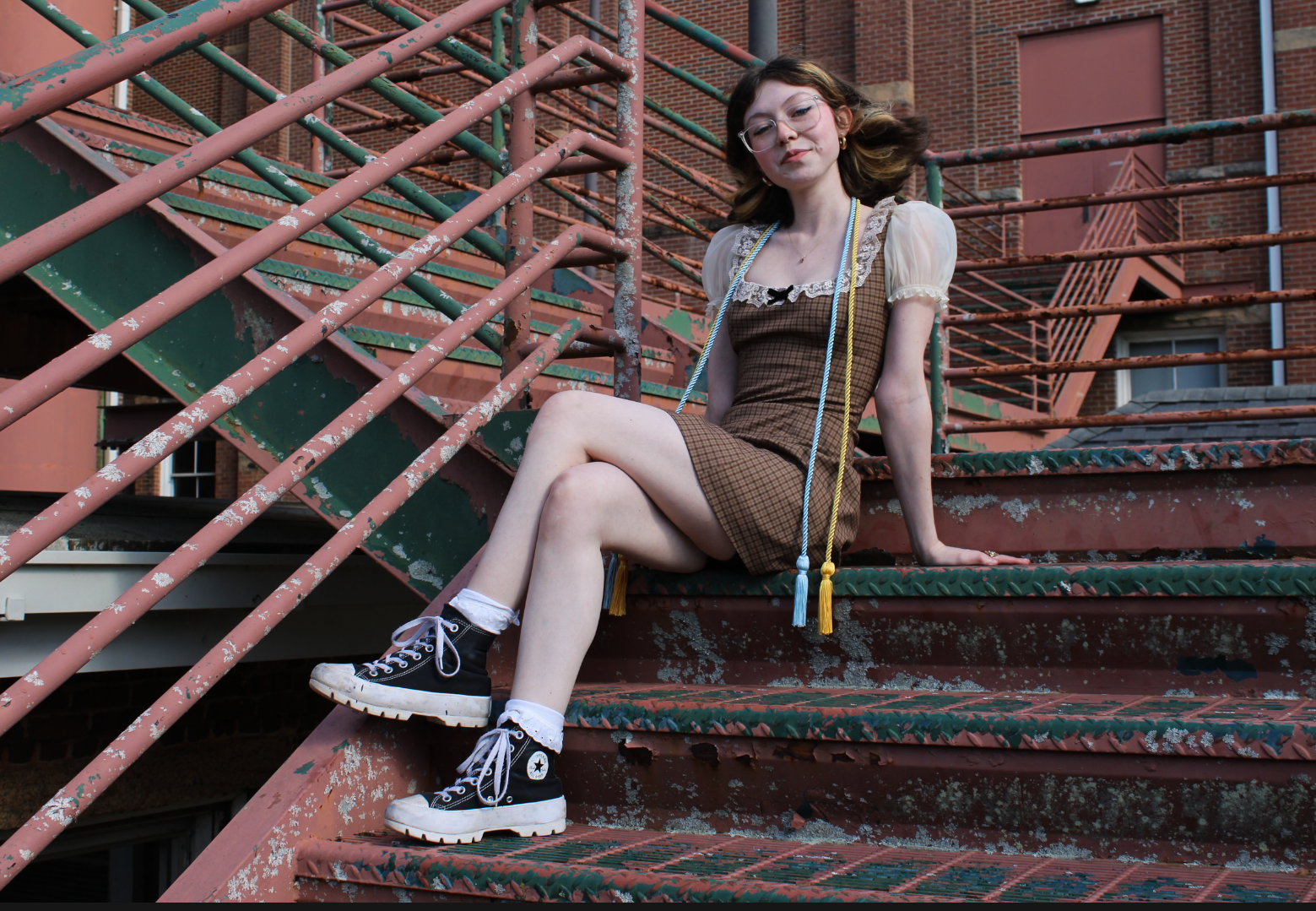 A young woman with glasses sitting on outdoor metal stairs painted in a rusty red color, wearing a plaid dress, black sneakers, and white socks, with a building in the background.