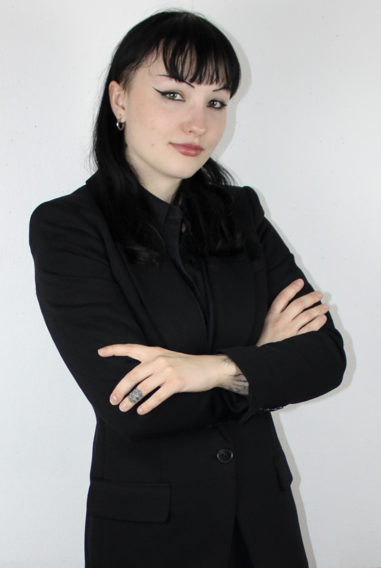 A young woman with black hair, wearing a black blazer, standing against a white wall with arms crossed and looking at the camera.