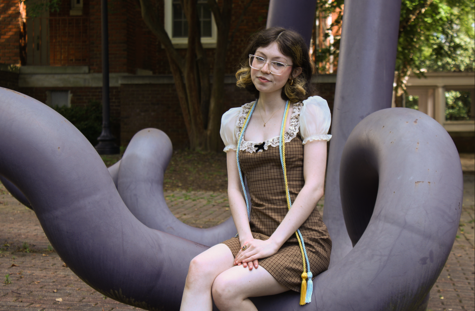 A young woman with glasses and shoulder-length wavy hair sits on a large, dark-colored sculpture in an outdoor park area. She is dressed in a vintage-style plaid dress with puffed sleeves and lace trim, and wears a graduation sash with a gold tassel.