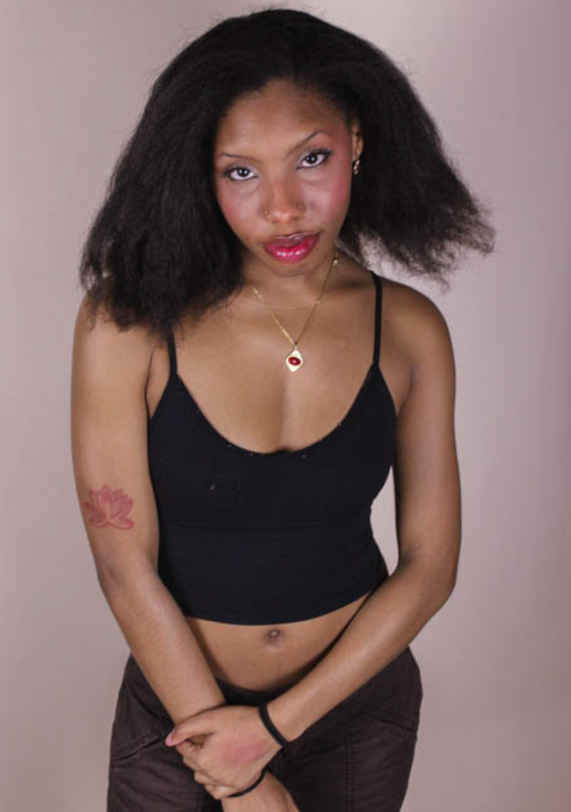 A young woman with dark, natural hair, wearing a black spaghetti strap top, dark pants, and a gold necklace with a red eye pendant. She has a pink tattoo on her left arm and is standing against a plain background, looking directly at the camera.