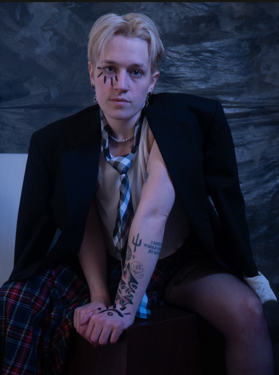 A person with short blonde hair and face paint, wearing a black blazer over a school uniform, sitting on a dark surface. They have tattoos on their arm and are posing with fists pressed together in front of them. The background is a dark, textured su