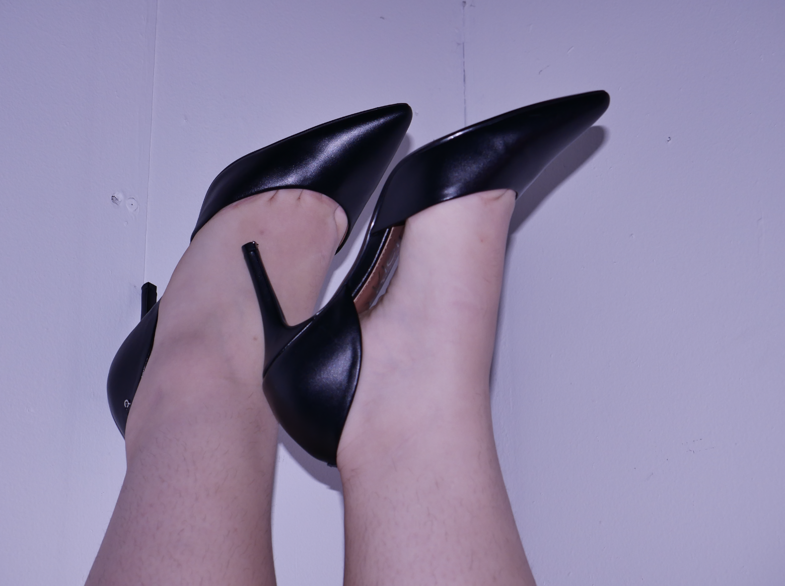 Close-up of a person's legs in black high heels with pointed toes, standing against a plain wall, viewed from below.