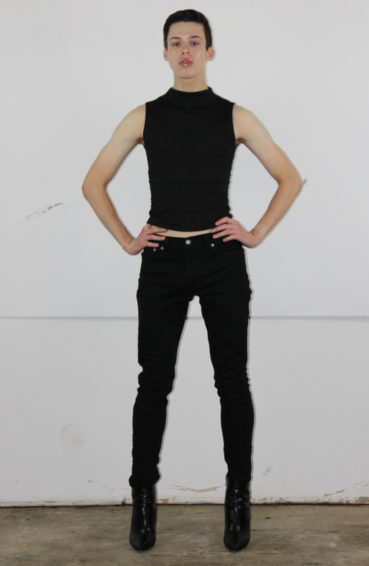 A young person stands with hands on hips, wearing a sleeveless black top, black pants, and black high-heeled boots, in front of a plain white wall.