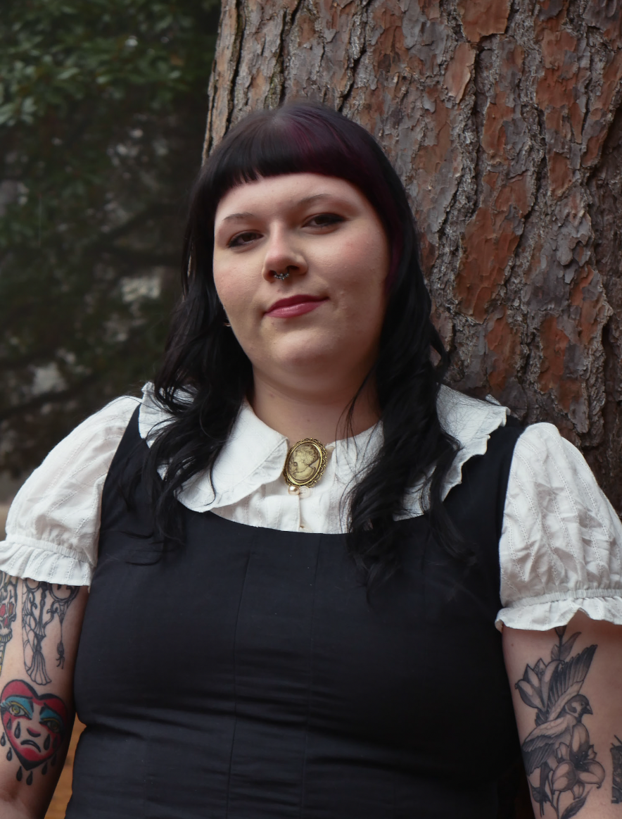 A woman with dark hair and tattoos, wearing a white blouse and black dress, standing outdoors next to a tree trunk.