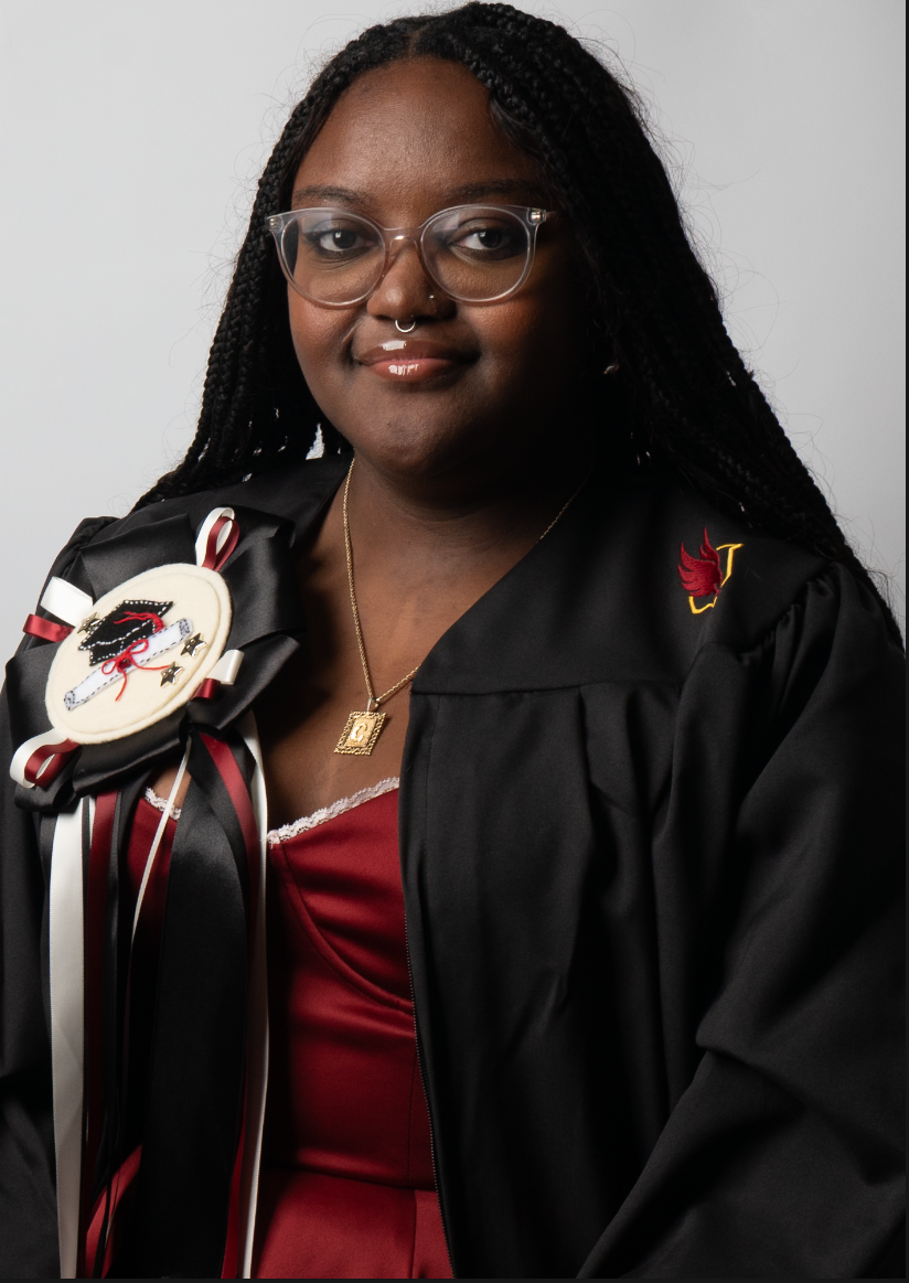 A young Black woman wearing graduation attire, including a black gown with a winged insignia on the sleeve, a red dress, glasses, and a hat pin with a black and white emblem. She has long braided hair, and is wearing jewelry including a necklace with