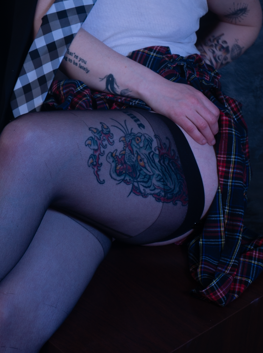 A person with tattoos, wearing fishnet stockings and a plaid skirt, sitting with their hand resting on their thigh. Visible tattoos include a dragon and a quote on their arm.