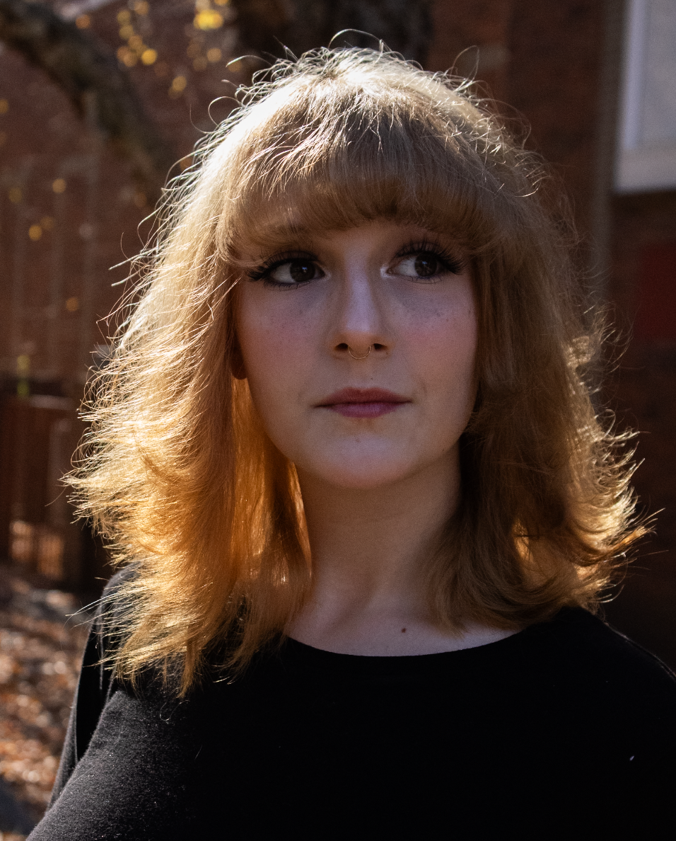 A young woman with shoulder-length wavy hair and a septum piercing outdoors during daylight, looking slightly to the side. She is wearing a black top and has natural makeup, with a background of a brick wall and greenery.