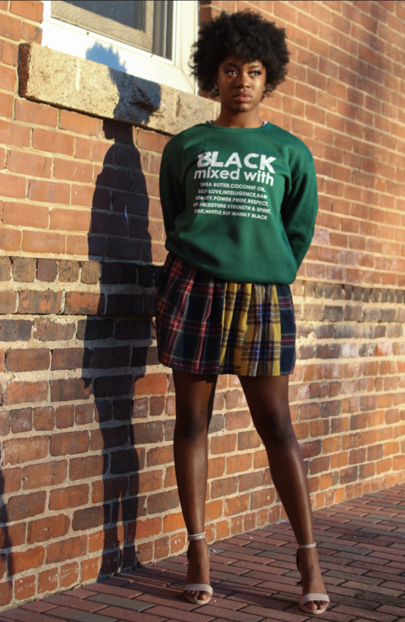 A young woman with curly hair standing against a brick wall, wearing a green sweatshirt with white text and a plaid skirt, with sunlight casting her shadow on the wall.