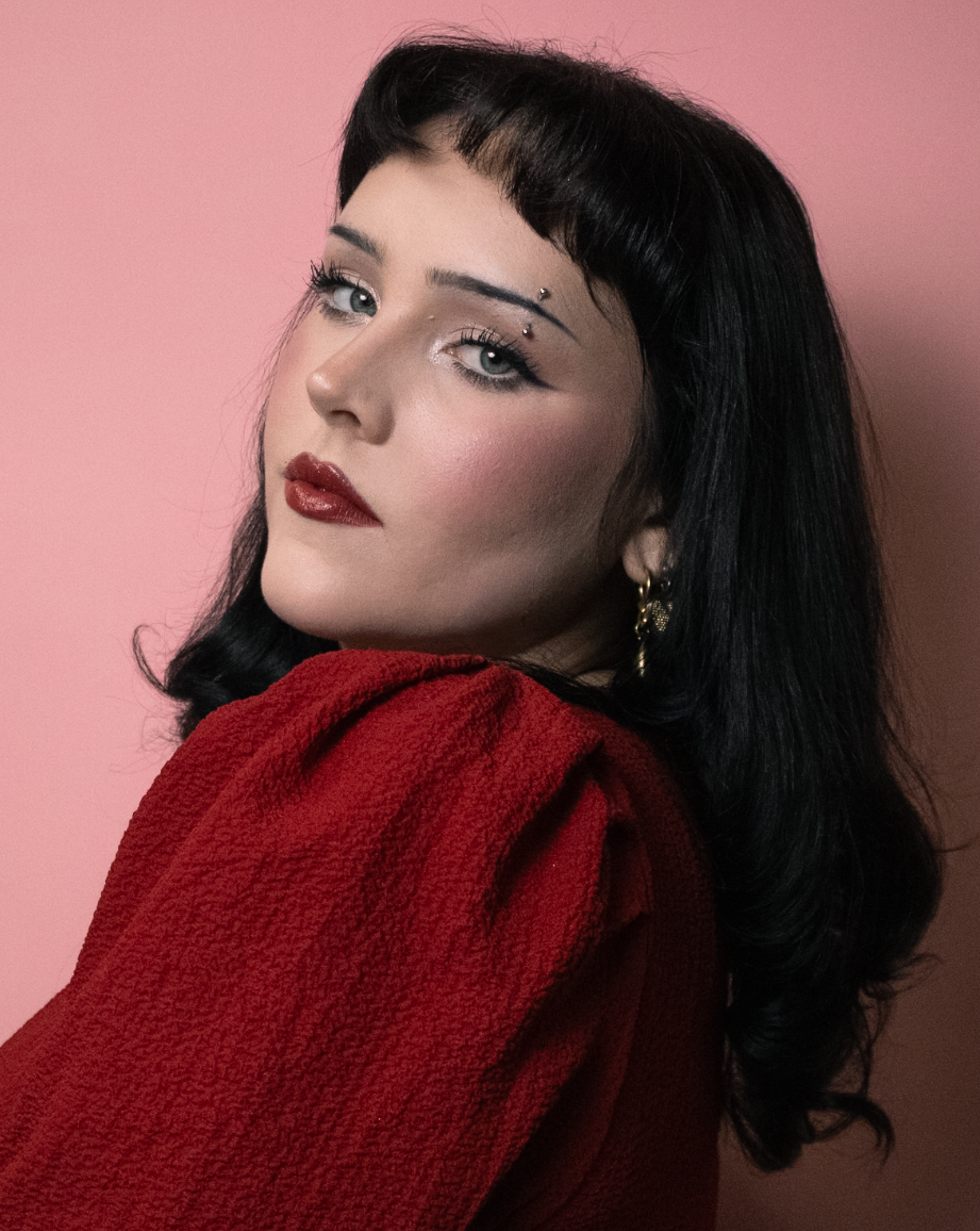 Portrait of a woman with black hair, wearing red lipstick, and a red textured top, against a pink background.