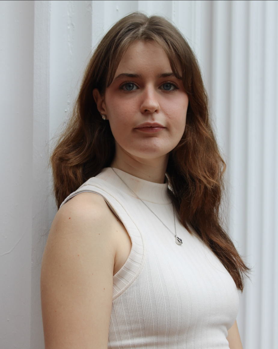 A young woman with shoulder-length wavy brown hair, fair skin, and blue eyes looking at the camera. She's wearing a sleeveless cream-colored top and a silver necklace with a heart-shaped pendant, standing in front of a white wall and vertical blinds.