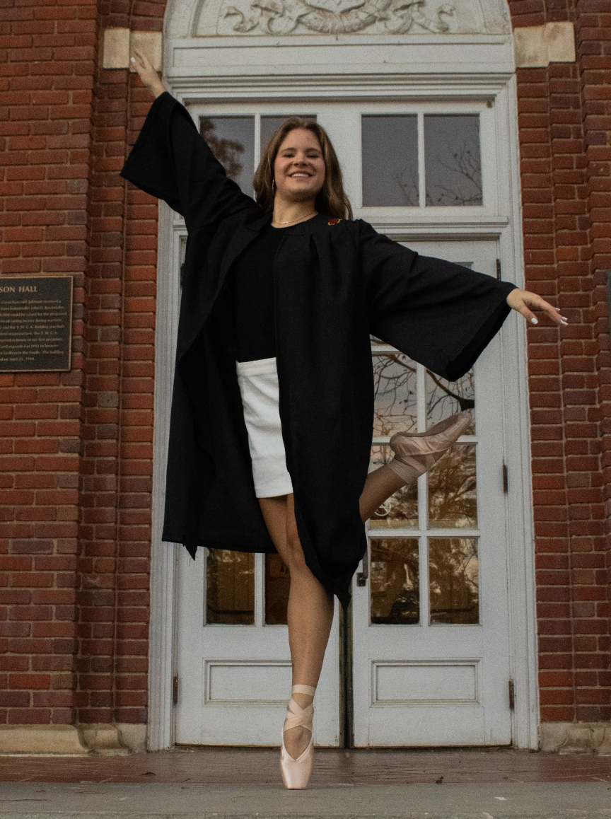 A young woman in a graduation gown and cap poses in a ballet stance on one tiptoe with her arms outstretched in front of a brick building with a white door.