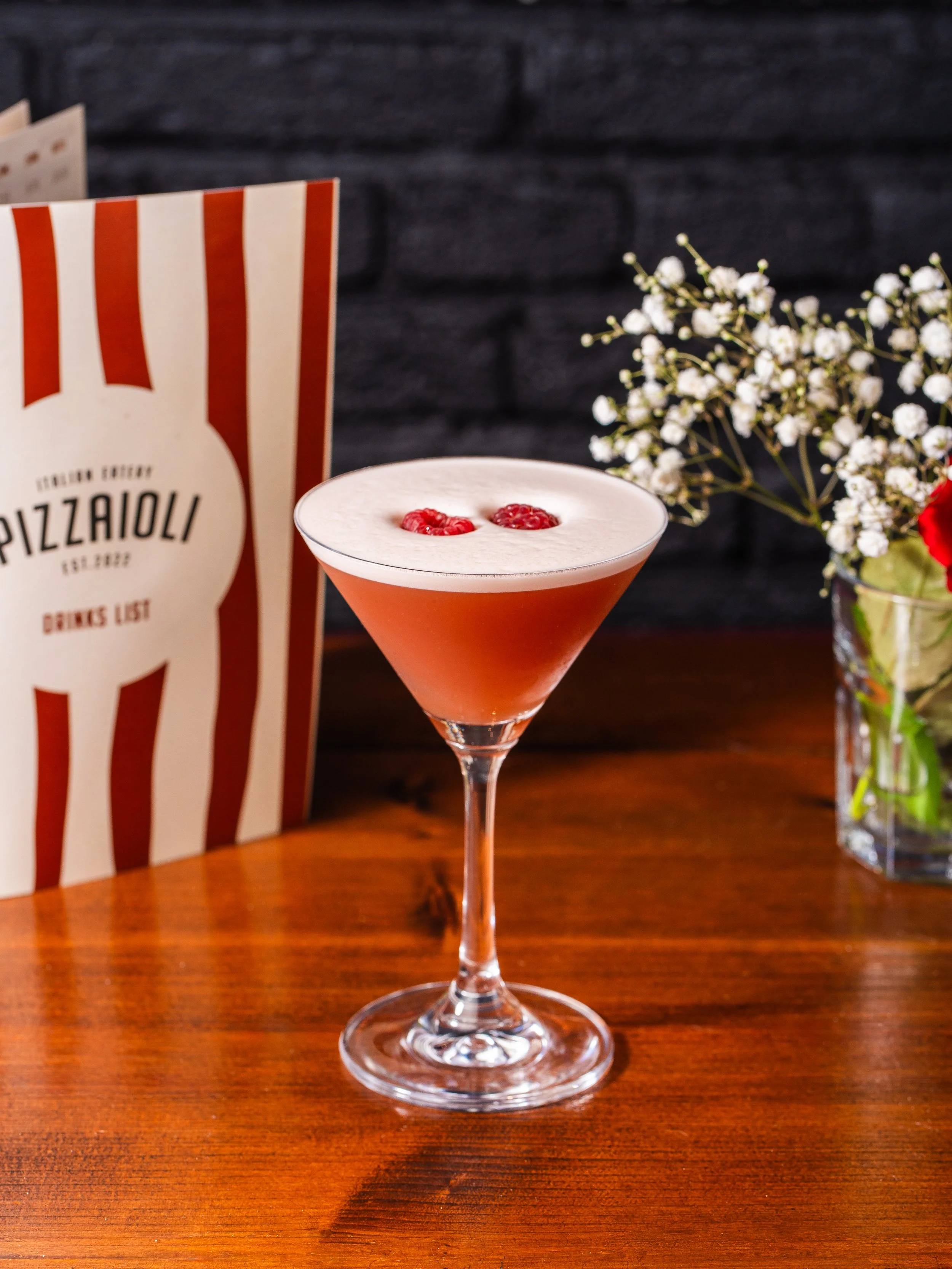 A pink cocktail with raspberries on top in a martini glass, positioned on a wooden table with a flower arrangement and a striped menu in the background.