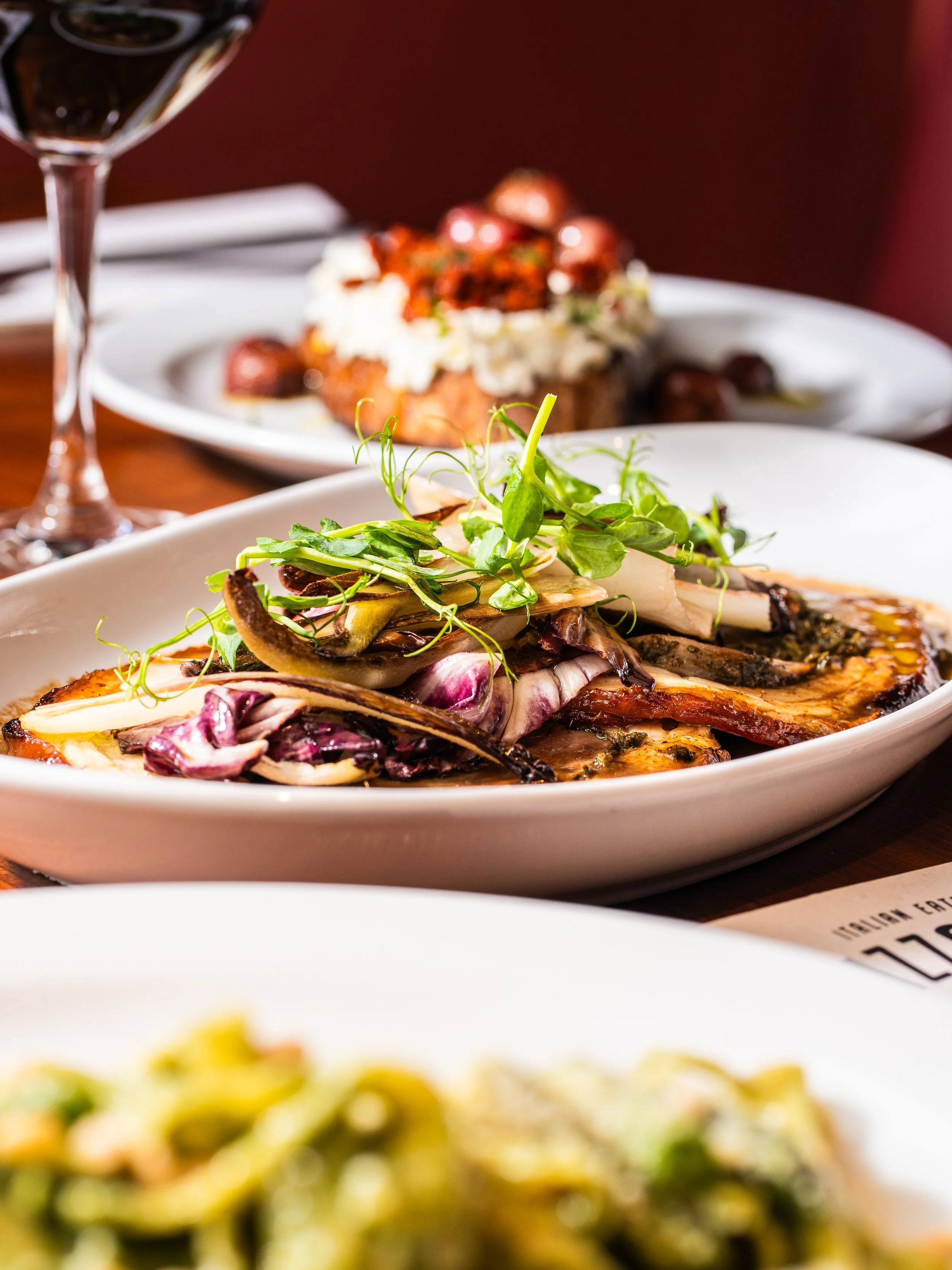 Close-up of a gourmet dish with grilled vegetables and microgreens in an oval white plate, with other dishes and a wine glass in the background.
