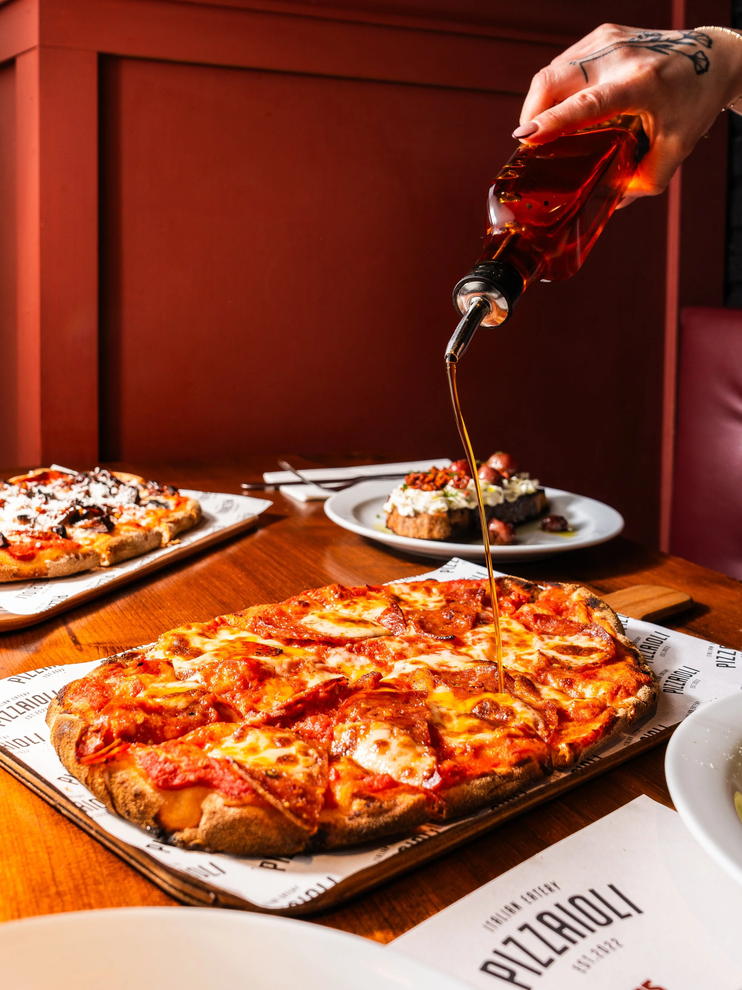 Hand pouring honey over a pepperoni pizza at a restaurant table with other pizzas and dishes.