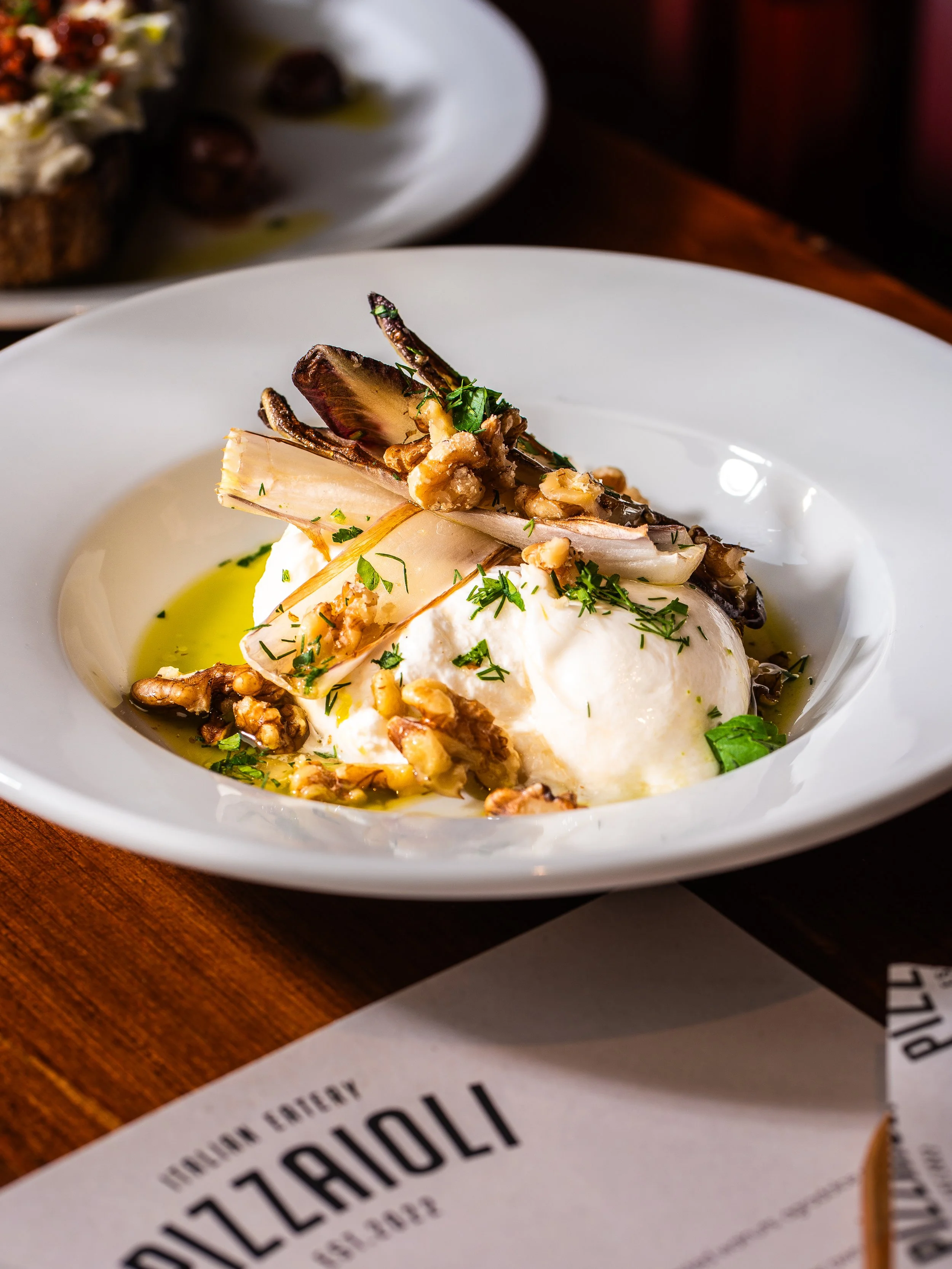 A white bowl containing burrata cheese topped with roasted vegetables, chopped herbs, and walnut pieces, drizzled with olive oil.