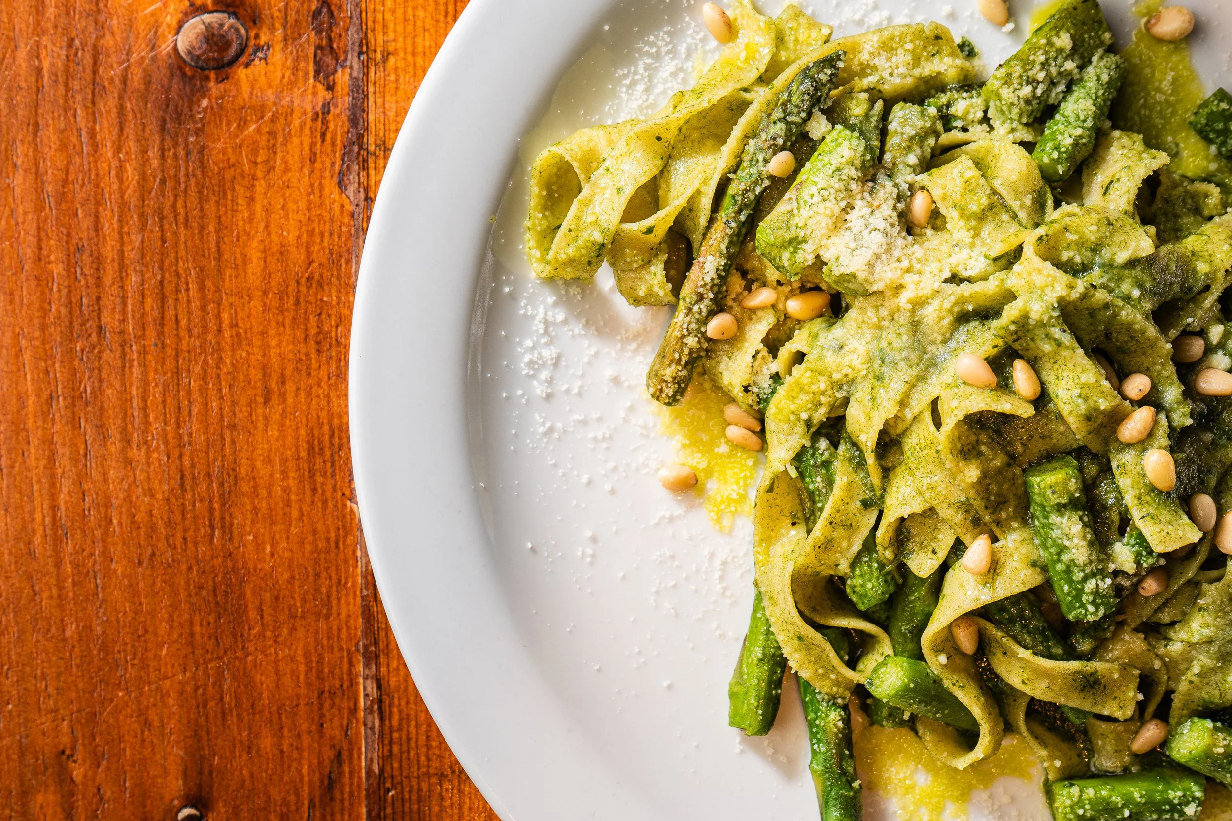 Close-up of a white plate with green vegetable pasta sprinkled with grated cheese and pine nuts, on a wooden surface.