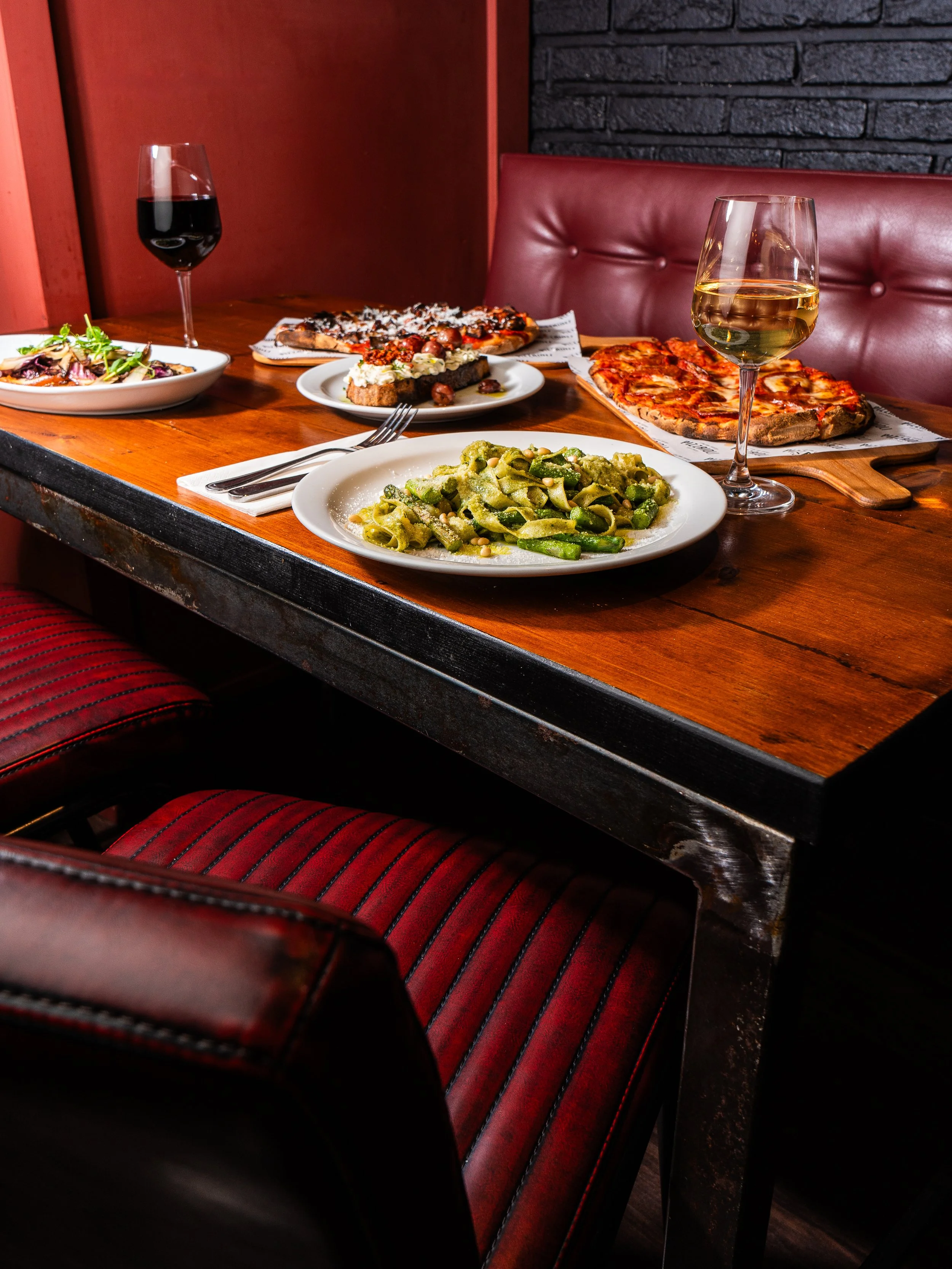 Table set with two glasses of wine, a pizza, pasta, salad, and dessert in a cozy restaurant.