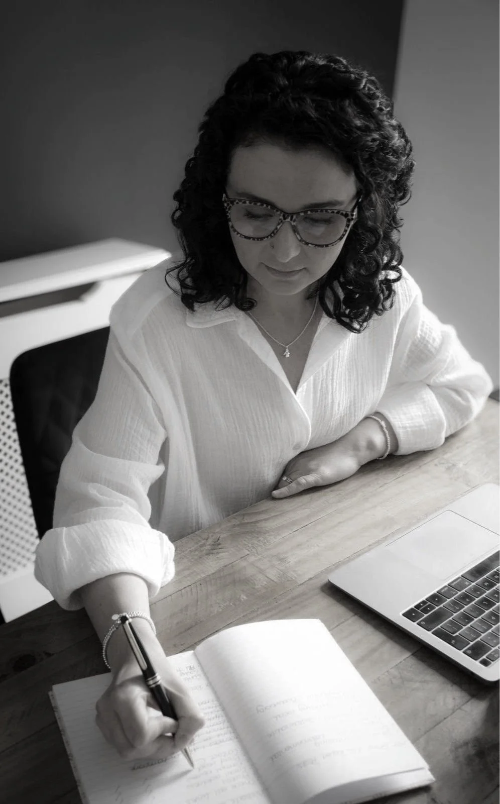 Woman with dark curly hair and glasses writing in notebook at desk with laptop