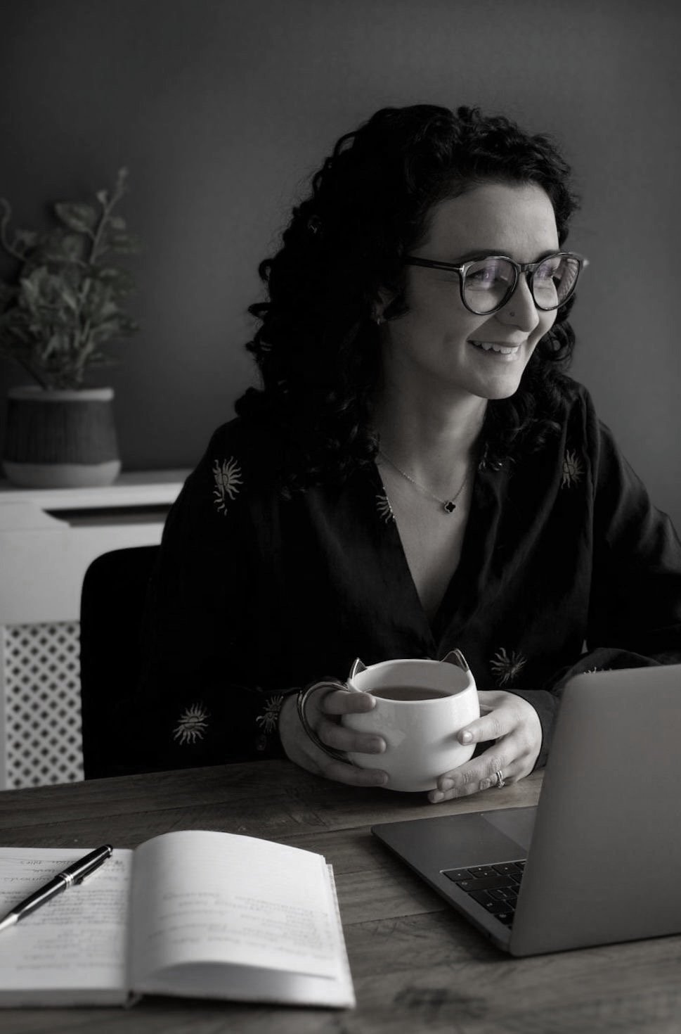 Woman with dark curly hair and glasses smiling while holding a coffee cup, sitting at a desk with a laptop, notebook, pen, and a plant nearby.